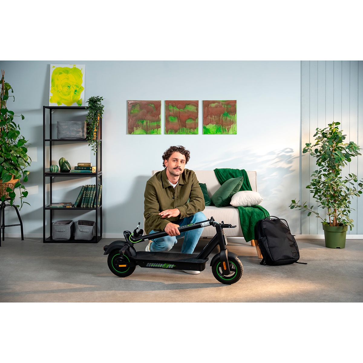 Hombre arrodillado junto a un patinete eléctrico en una sala con arte y plantas.