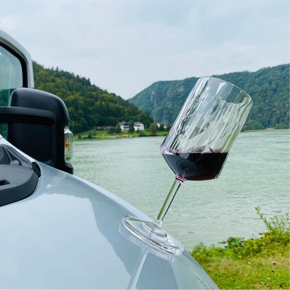 Un verre de vin rouge repose sur un véhicule argenté, avec un lac et des collines vertes en arrière-plan.