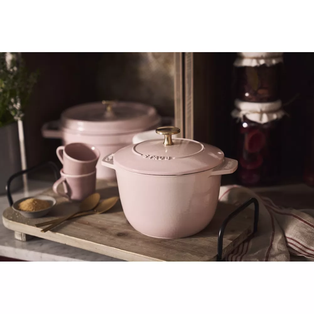 Cocotte en fonte rose avec poignée dorée sur plateau en bois, près de tasses.