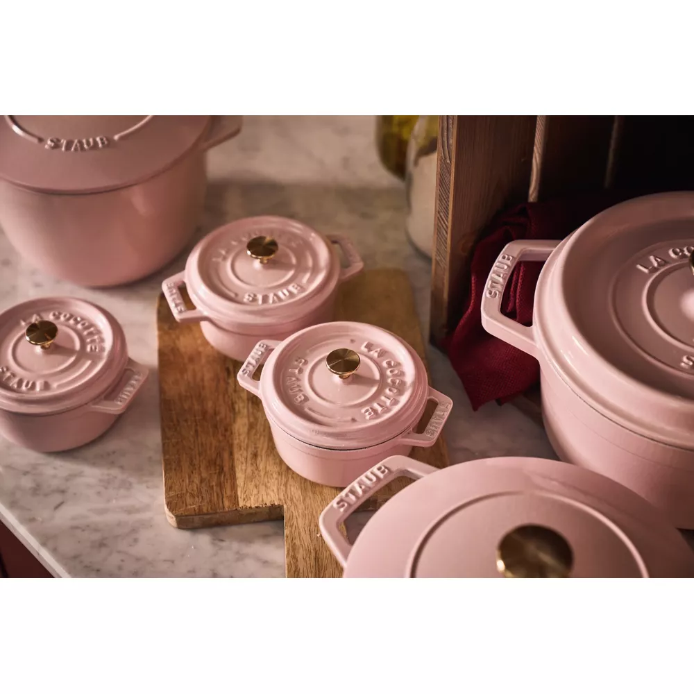 Plusieurs ustensiles de cuisine Staub roses à boutons dorés sur une surface en bois.