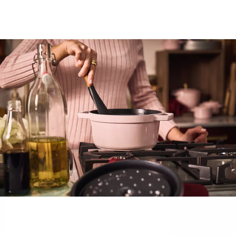 Une personne remue une cocotte rose pâle sur une cuisinière à gaz. Des bouteilles d'huile et de sauce sont à proximité.