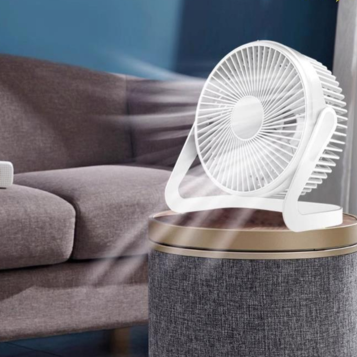 Ein weißer Ventilator auf einem grauen Hocker, der Luft auf ein Sofa bläst.