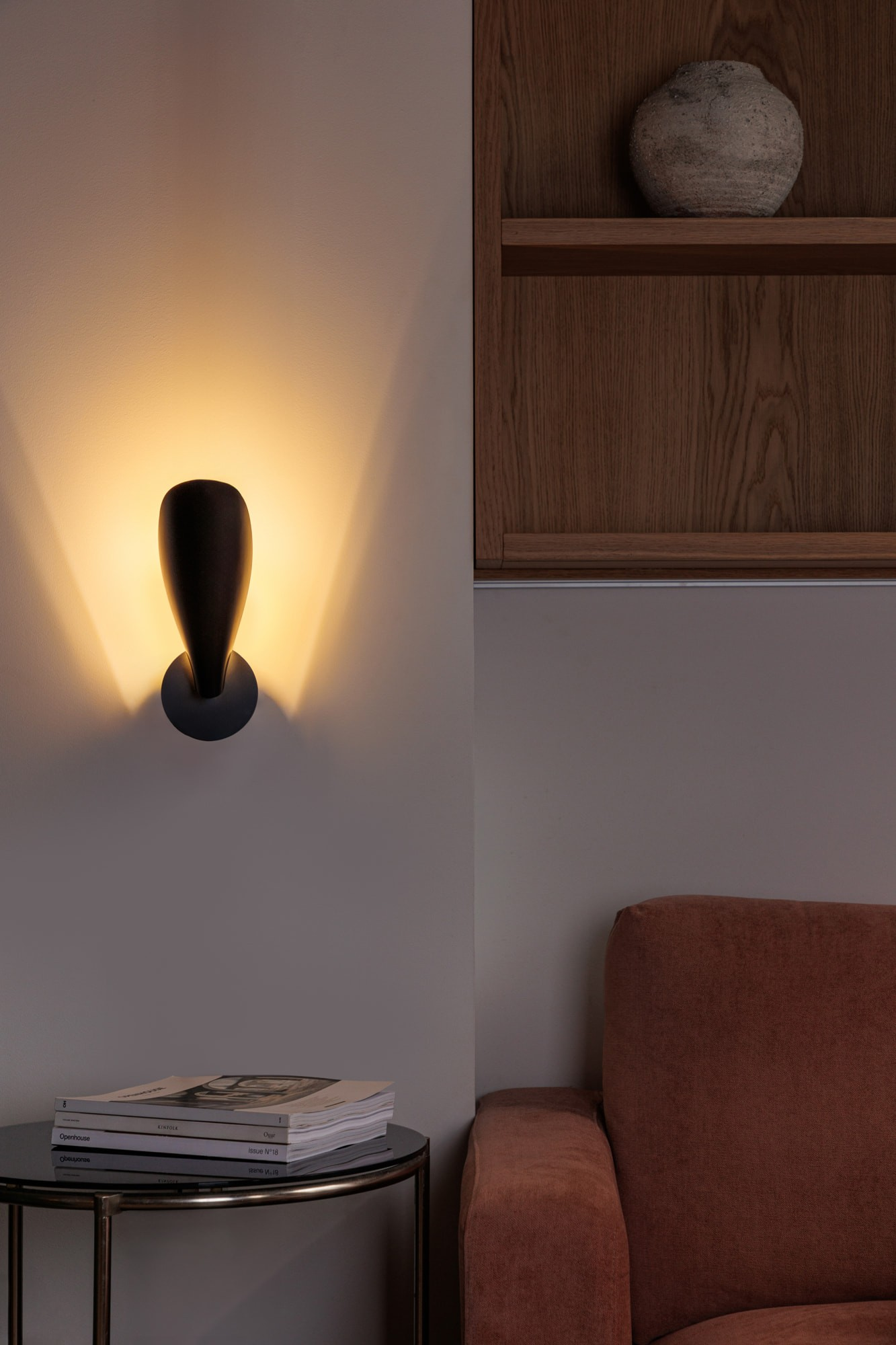 Una lampada da parete proietta luce calda su un muro accanto a una mensola in legno con vaso.