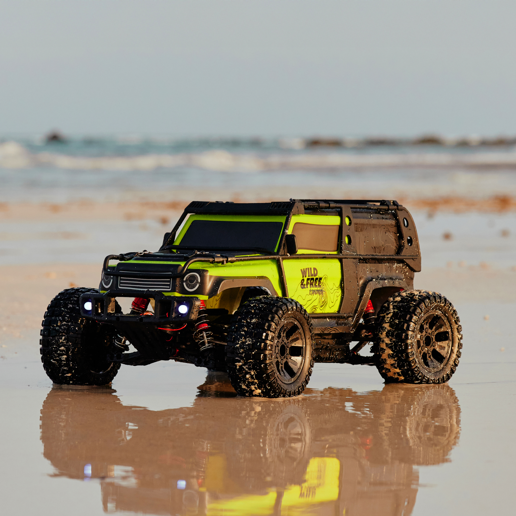 Ein gelbes RC-Auto mit großen Reifen steht am nassen Sandstrand, mit dem Meer im Hintergrund.