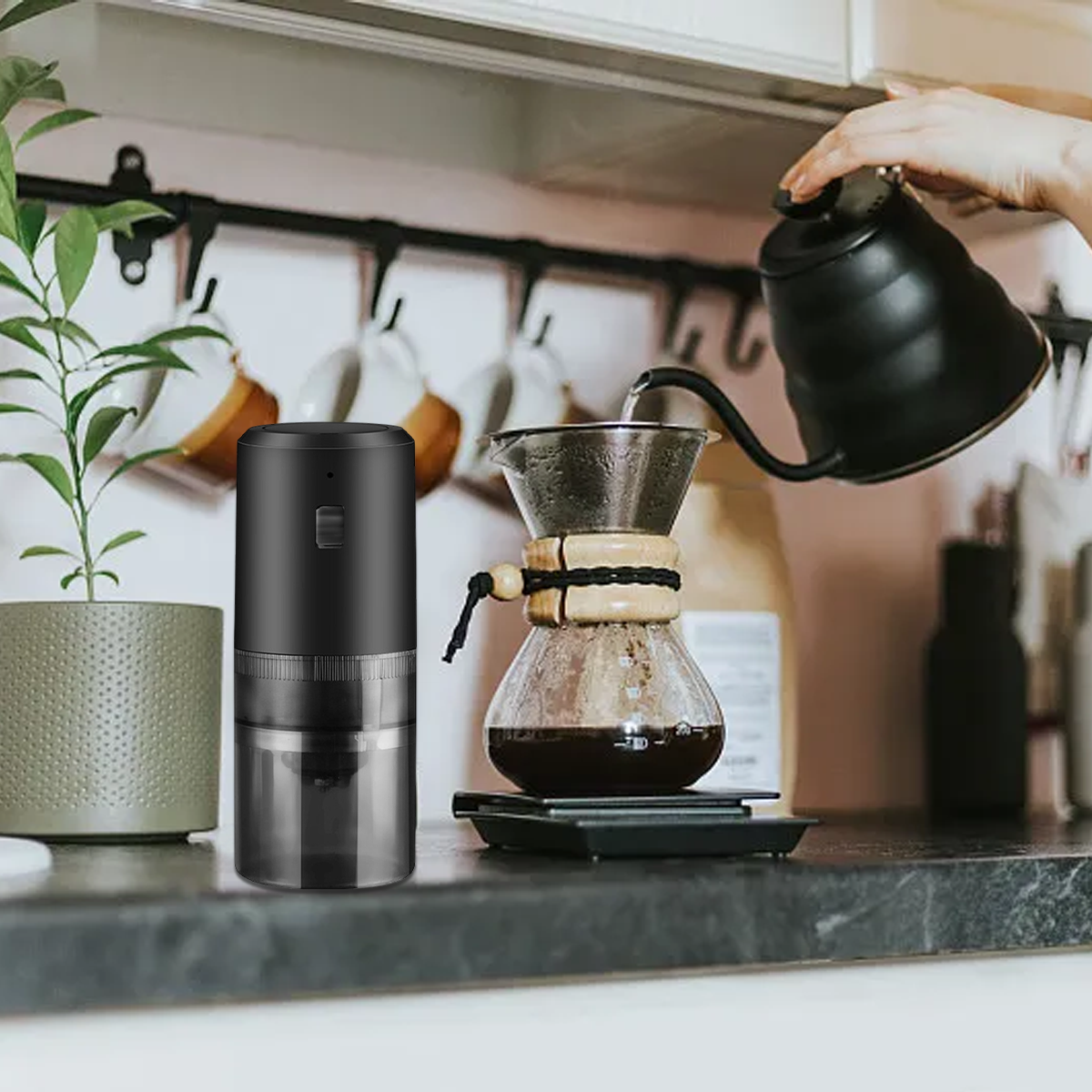Eine schwarze Kaffeemühle und ein Chemex-Brüher stehen auf einer Theke. Wasser wird aus einem Wasserkocher gegossen.
