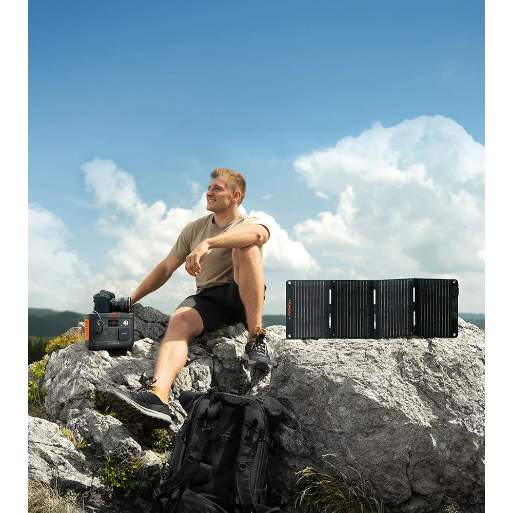 Uomo seduto su rocce vicino a pannello solare e power station, zaino davanti.