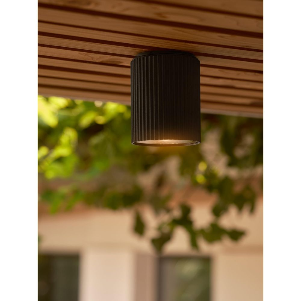 Lampada cilindrica nera scanalata su soffitto in legno, fogliame verde dietro.