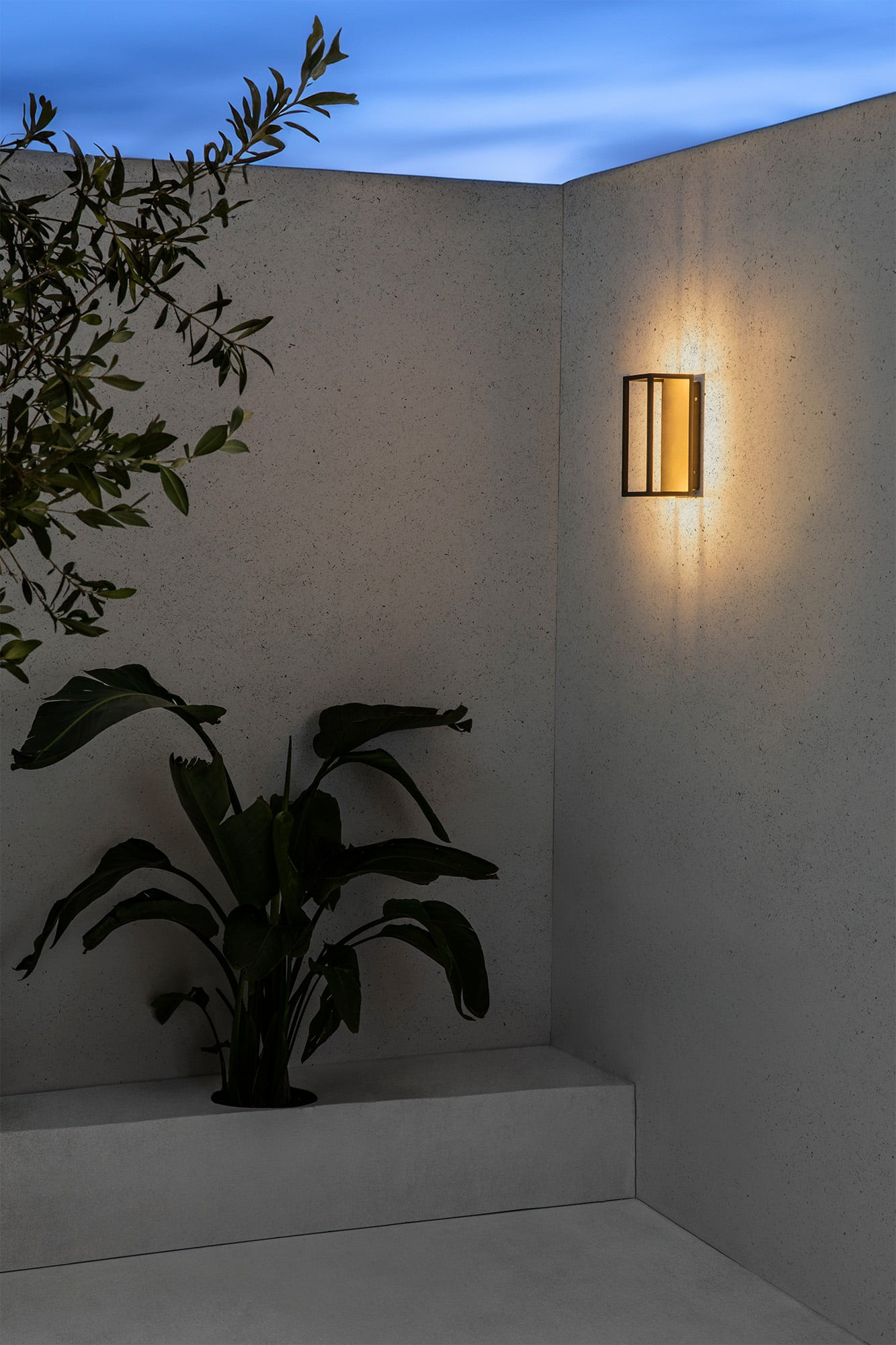 Lampada da parete esterna illumina muro texturizzato vicino a piante verdi sotto cielo crepuscolare.