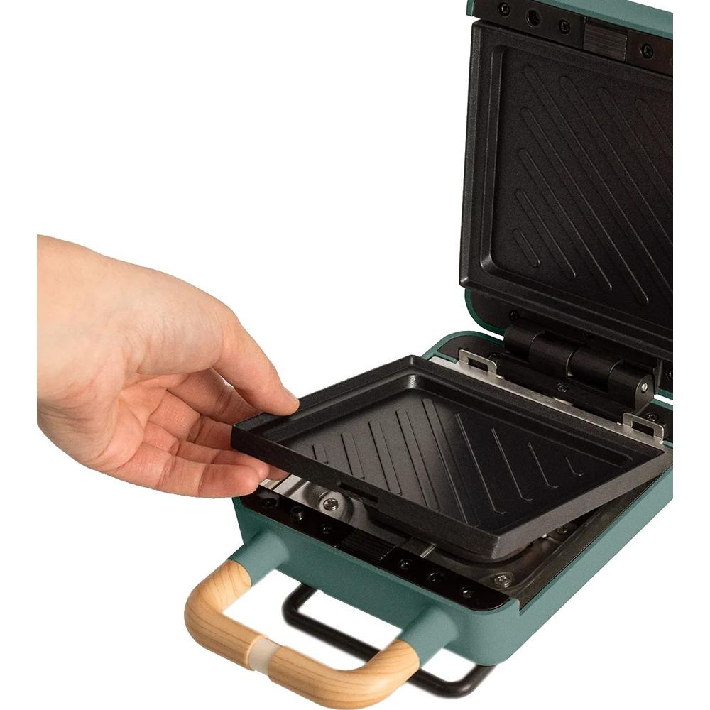 Una mano inserta una plancha en una sandwichera verde con asas de madera.