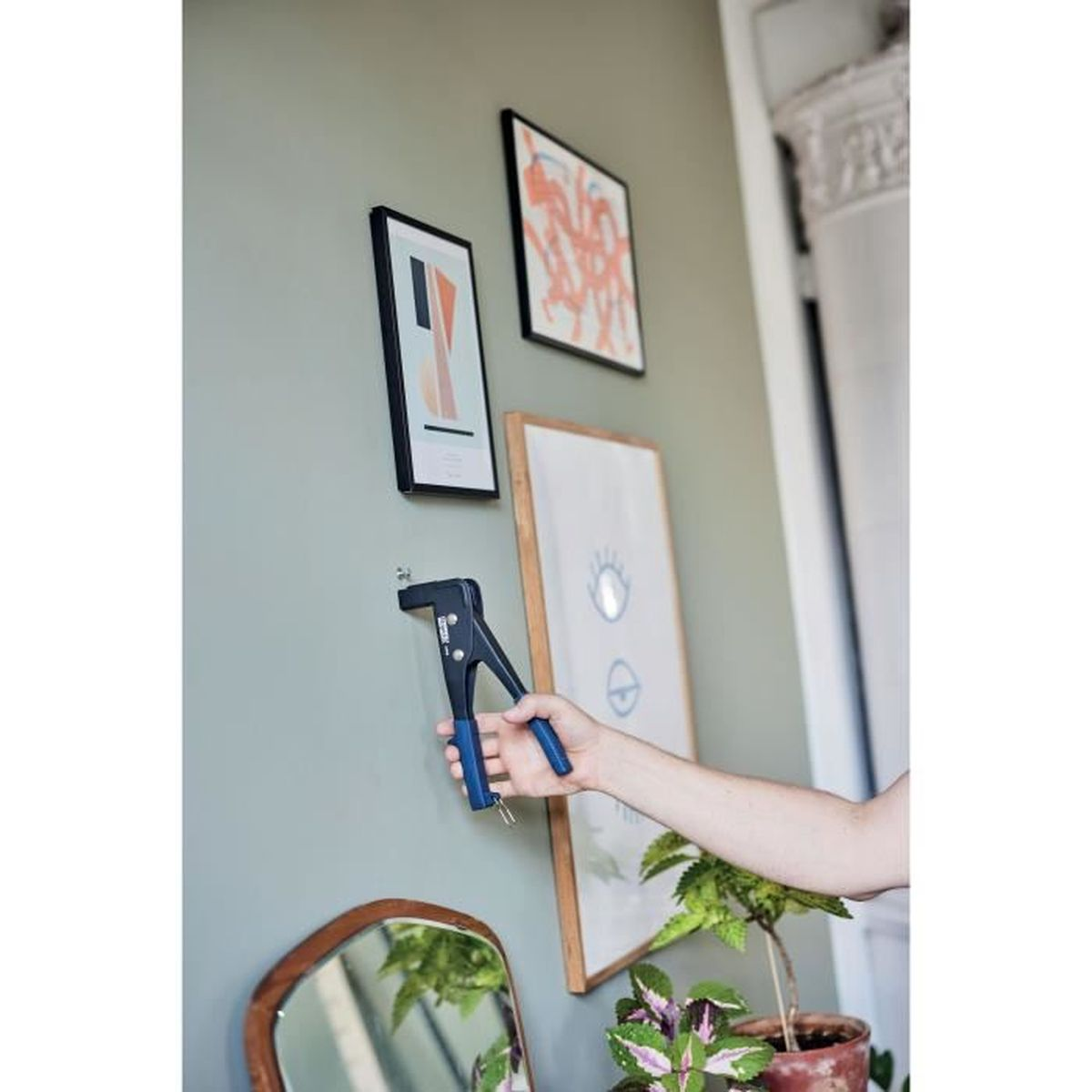 Eine Hand benutzt eine Zange, um einen Haken an einer Wand zu befestigen. Mehrere Bilder hängen an der Wand.