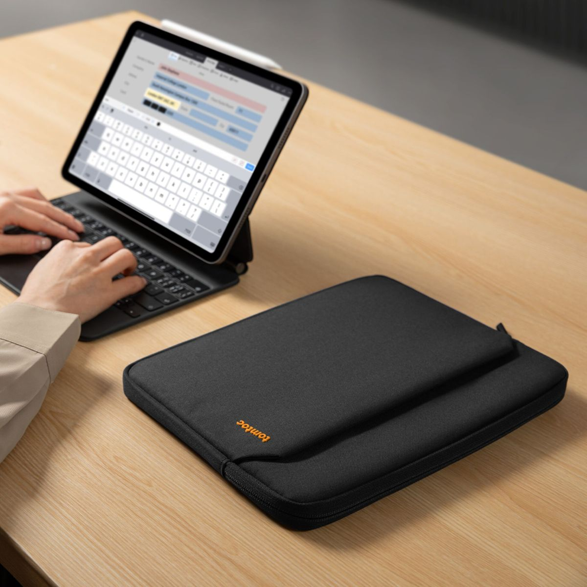 Eine Person tippt auf einer Tastatur, ein Tablet mit Stift und eine schwarze Laptoptasche liegen auf einem Holztisch.