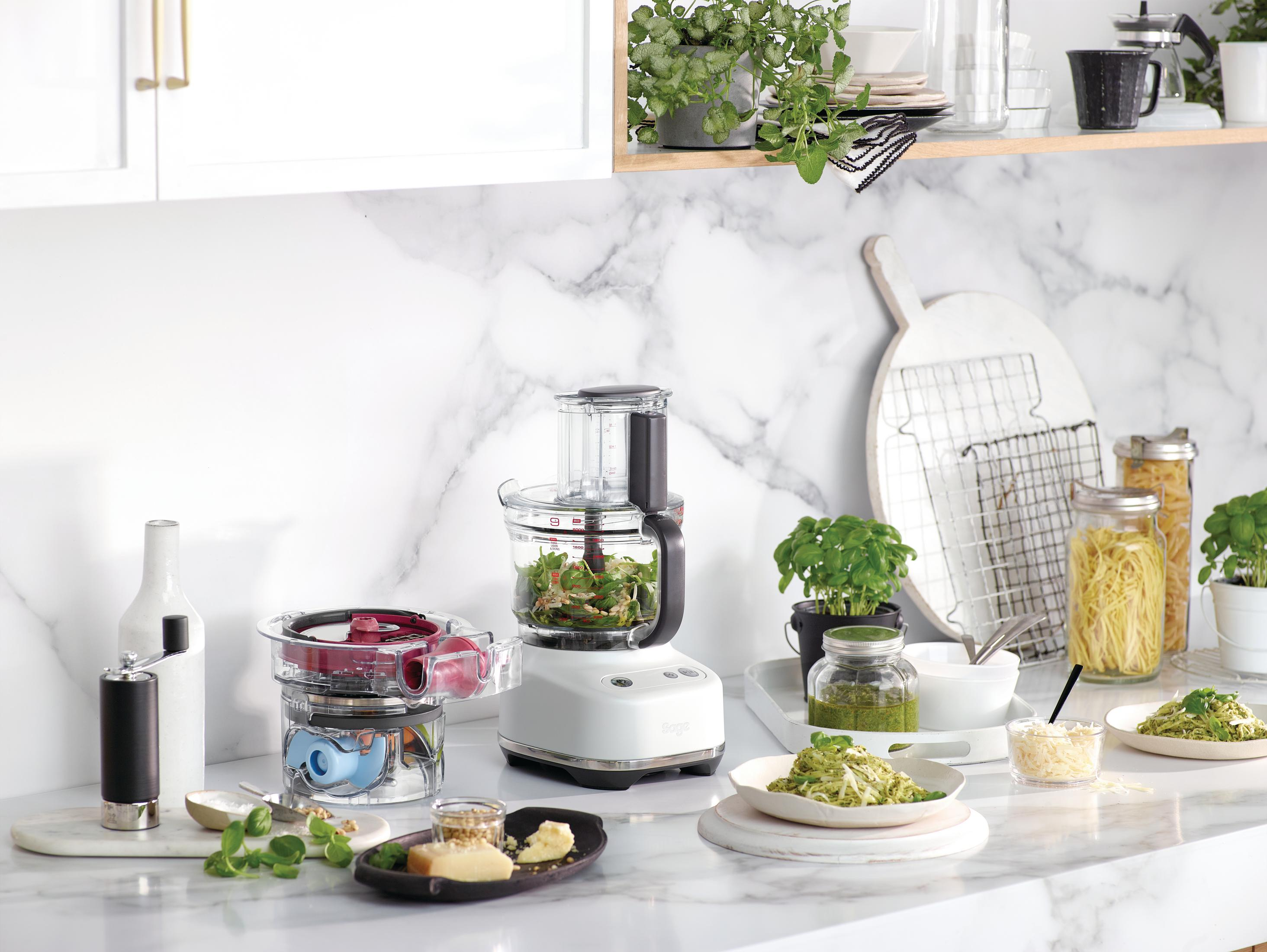 Eine weiße Küchenmaschine mit Zutaten für Pesto, Nudeln und Käse.