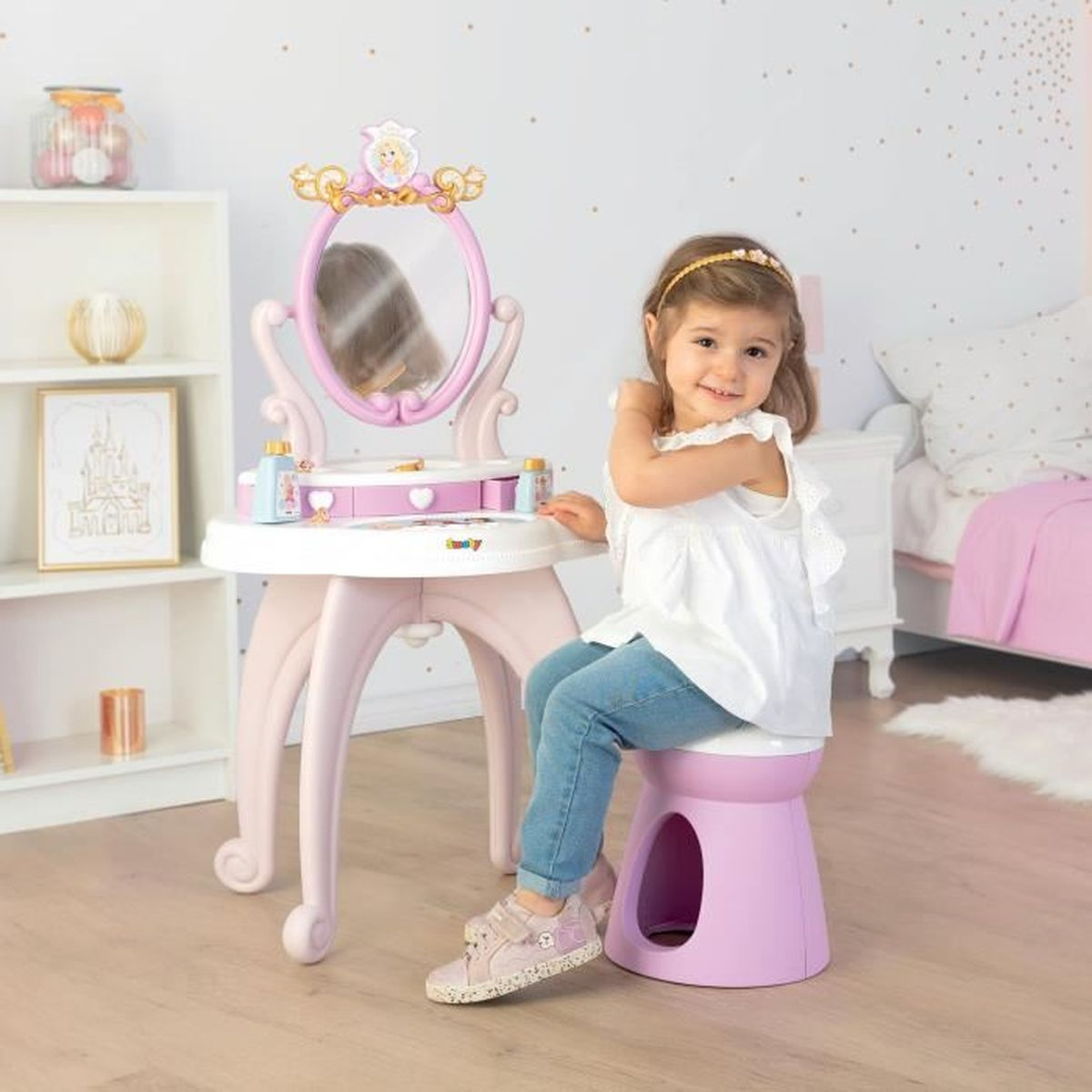 Una bambina siede a un tavolino da trucco con specchio. Indossa un top bianco e jeans.