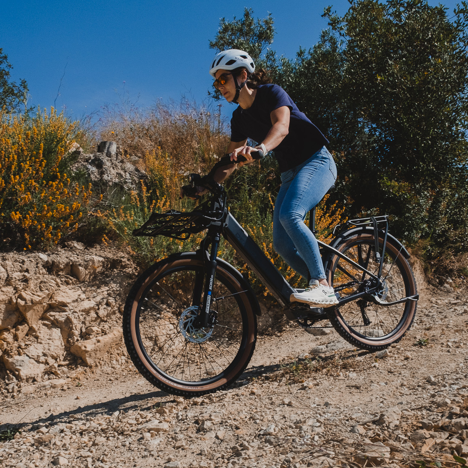Frau mit Helm fährt E-Bike auf Feldweg mit Bäumen und gelben Blumen.