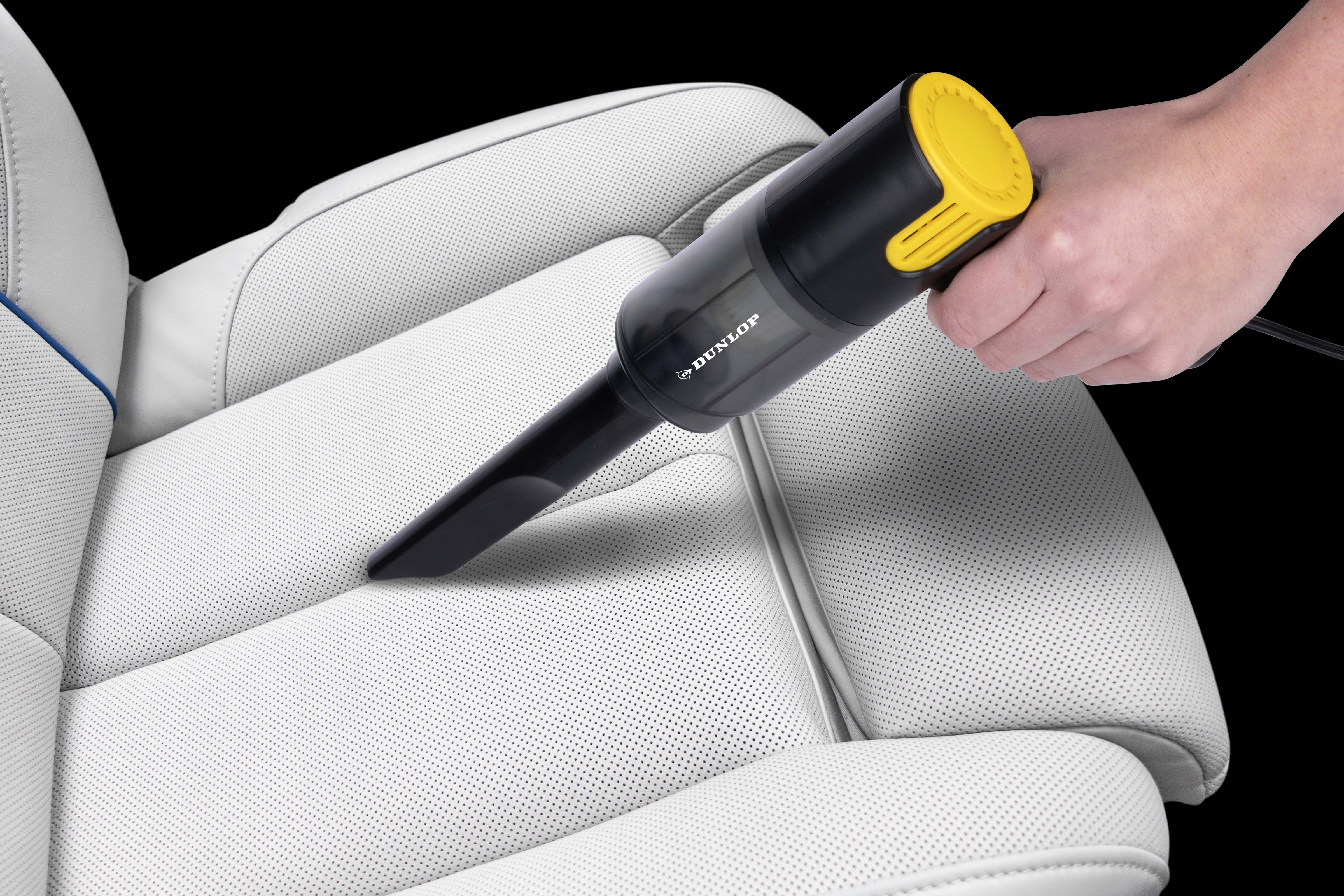 A hand uses a black and yellow handheld vacuum on a white leather car seat.
