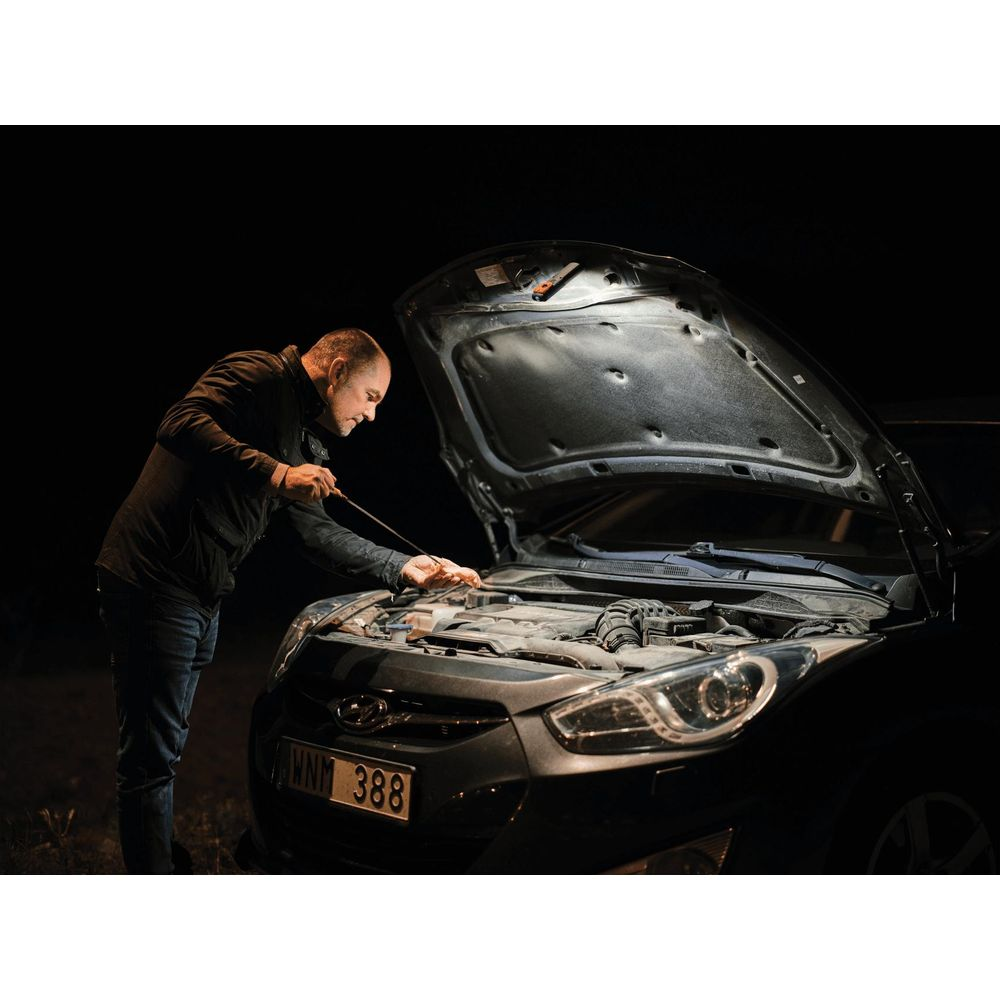 Un homme vérifie le moteur d'une voiture la nuit, capot ouvert.