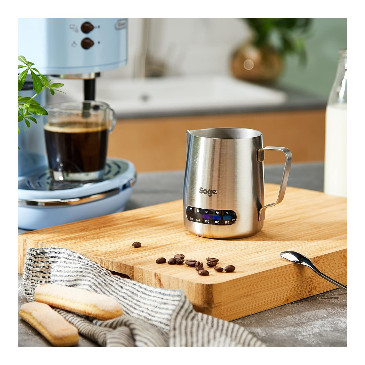 A silver milk frothing pitcher with a thermometer sits on a wooden board. Coffee machine in background.