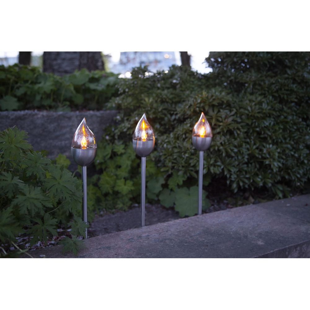 Drei flammenförmige Solarleuchten stehen im Garten. Sie haben silberne Stiele und Glasköpfe.