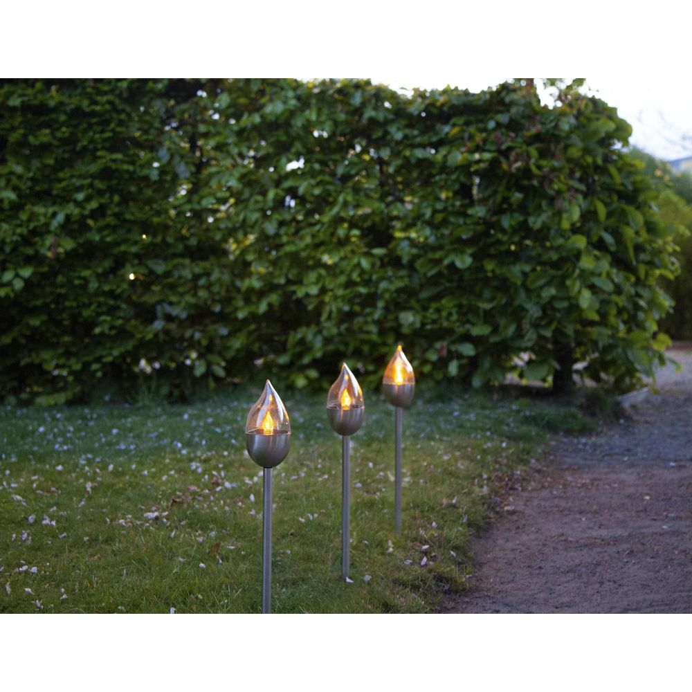Drei Flammen-Gartenleuchten stehen auf einem Rasen vor einer dichten Hecke.