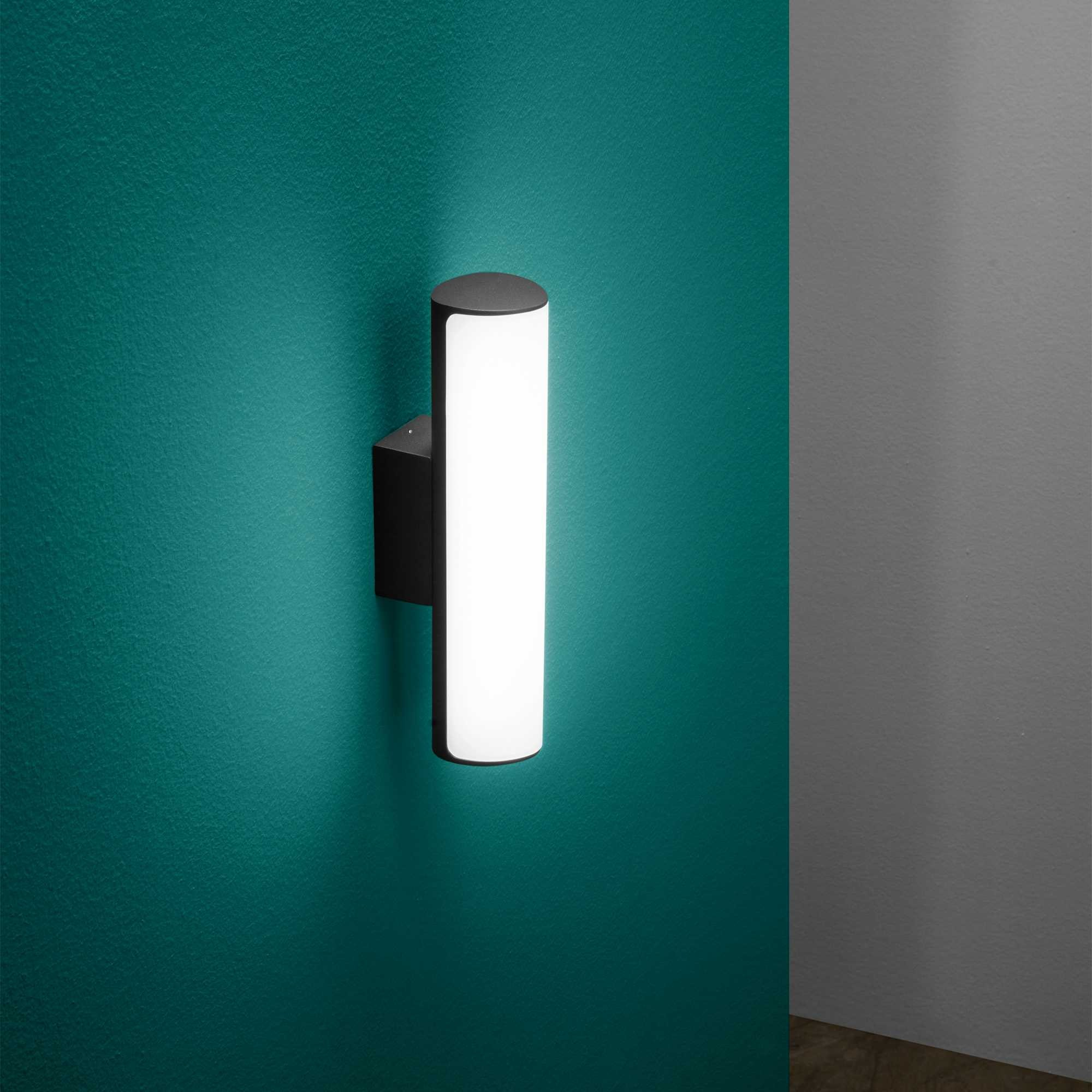 Una lampada da parete cilindrica con base nera illumina una parete verde.