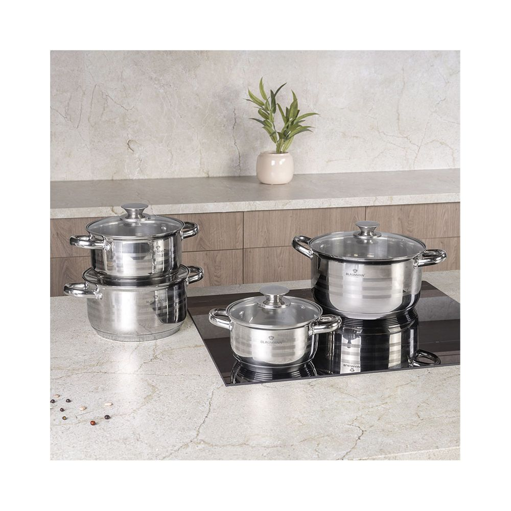 Quatre casseroles en acier inoxydable avec couvercles en verre sur une table de cuisson. Une plante à l'arrière.