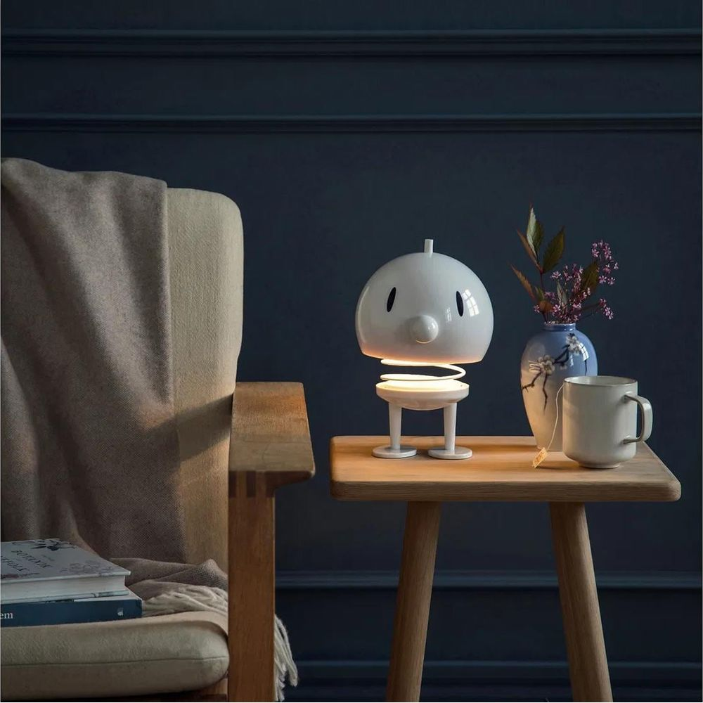Une lampe blanche de style dessin animé est sur une table en bois à côté d'un vase et d'une tasse.