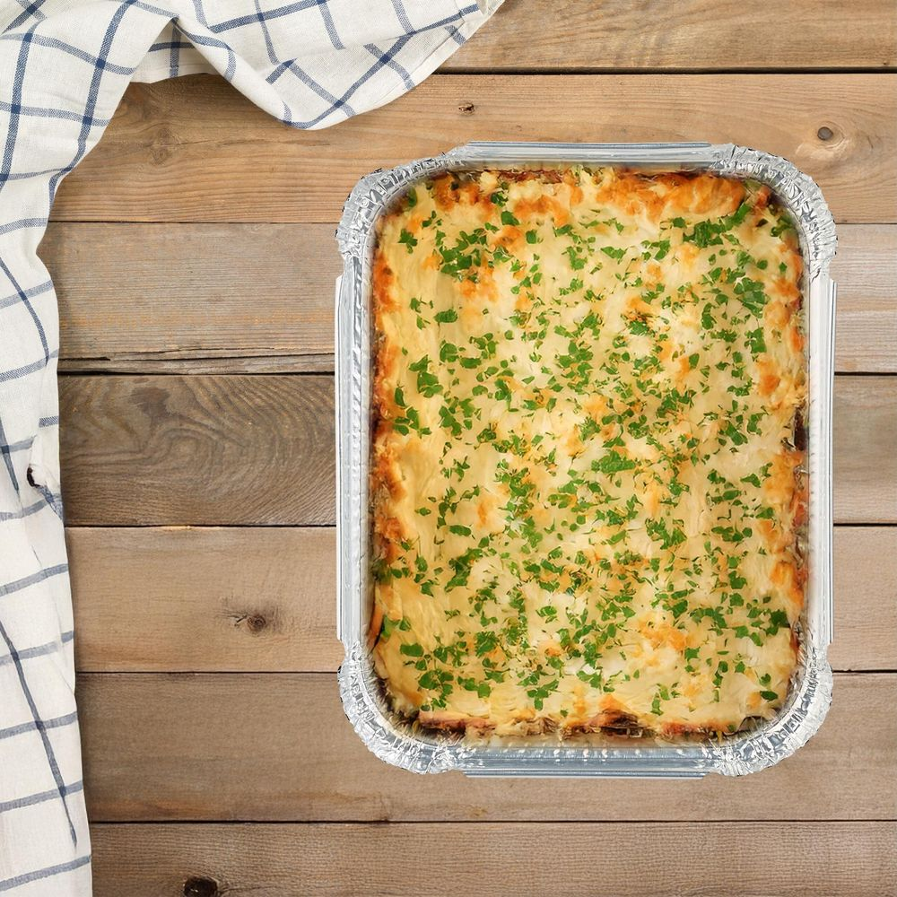 Lasagna in teglia di alluminio, guarnita con prezzemolo su tavolo di legno.