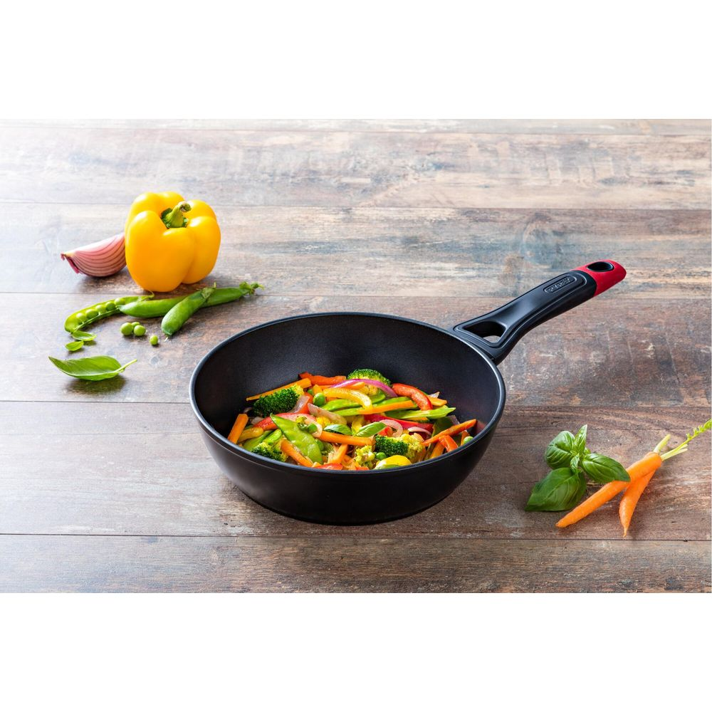 Un wok noir rempli de légumes colorés est sur une table en bois. Ingrédients à proximité.