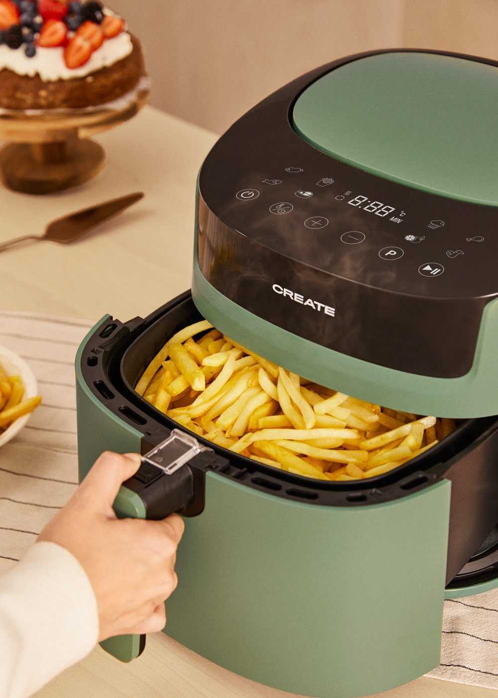 Una friggitrice ad aria verde piena di patatine fritte, con una torta vicina.