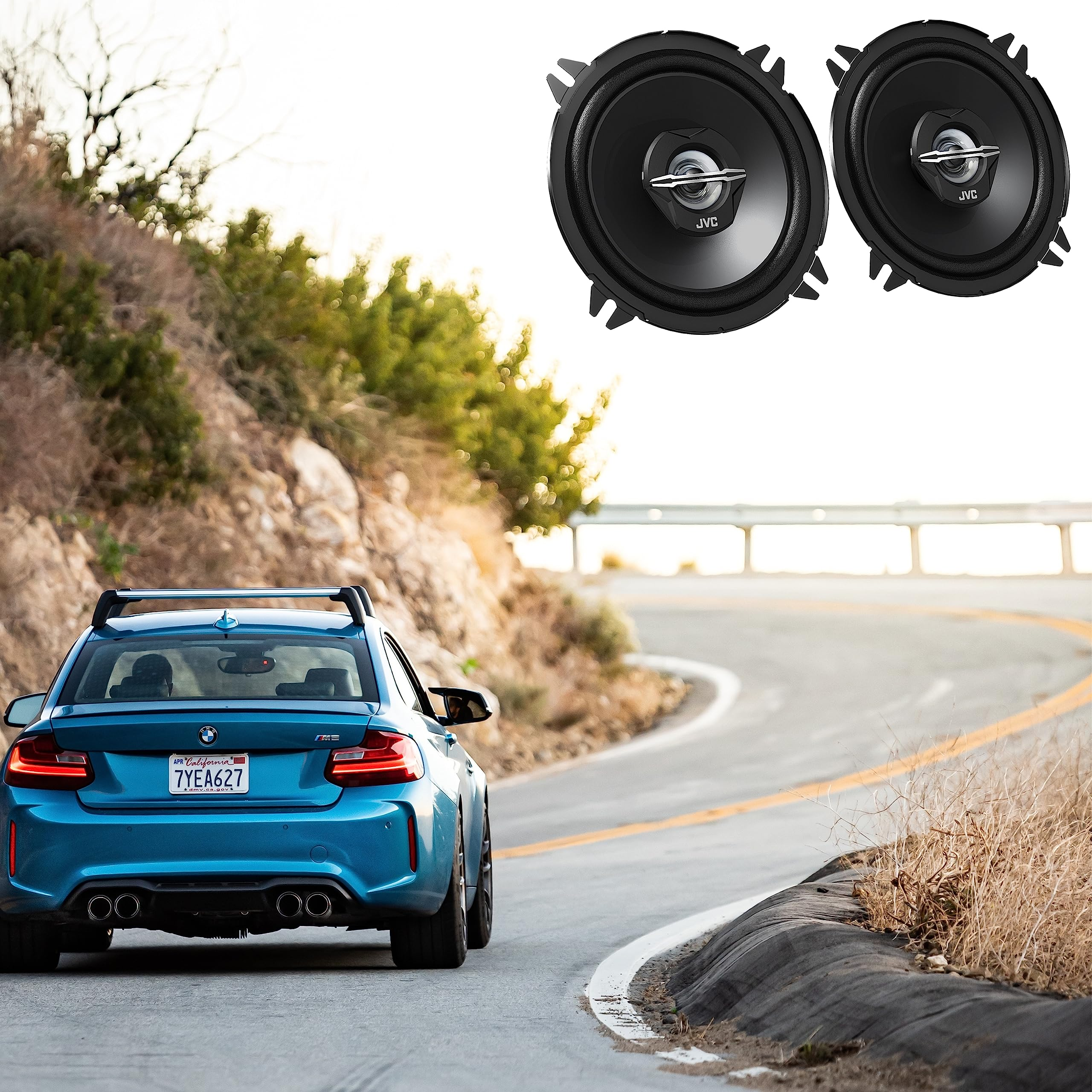 Zwei JVC-Lautsprecher über einem blauen BMW auf einer kurvigen Straße.