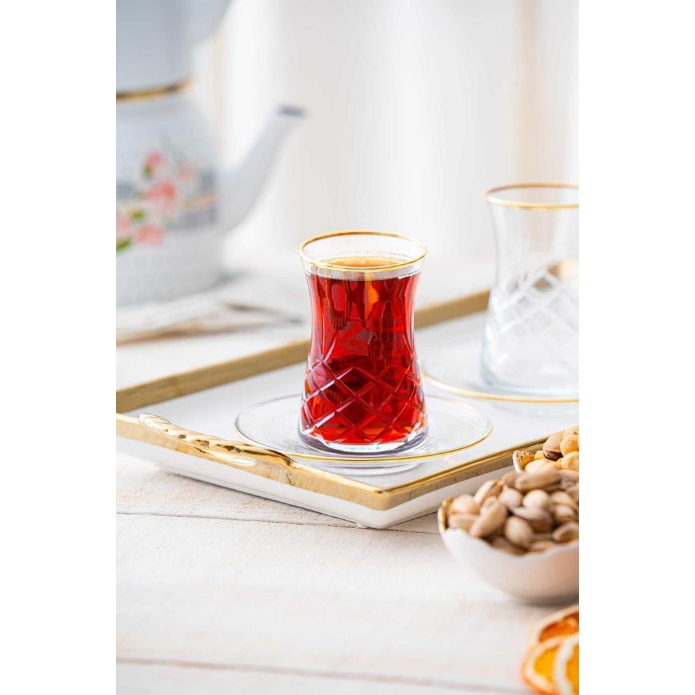 Altın kenarlı koyu çay bardağı tepside bir fincan tabağında duruyor. Arkada çaydanlık ve başka bir bardak var.