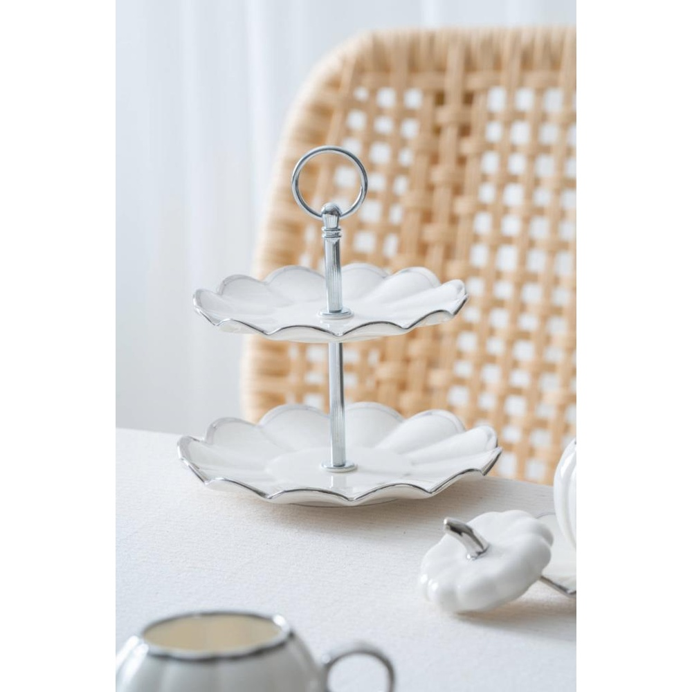 Gümüş kenarlı iki katlı beyaz servis standı beyaz masa örtüsü üzerinde duruyor. Arkasında hasır sandalye var.