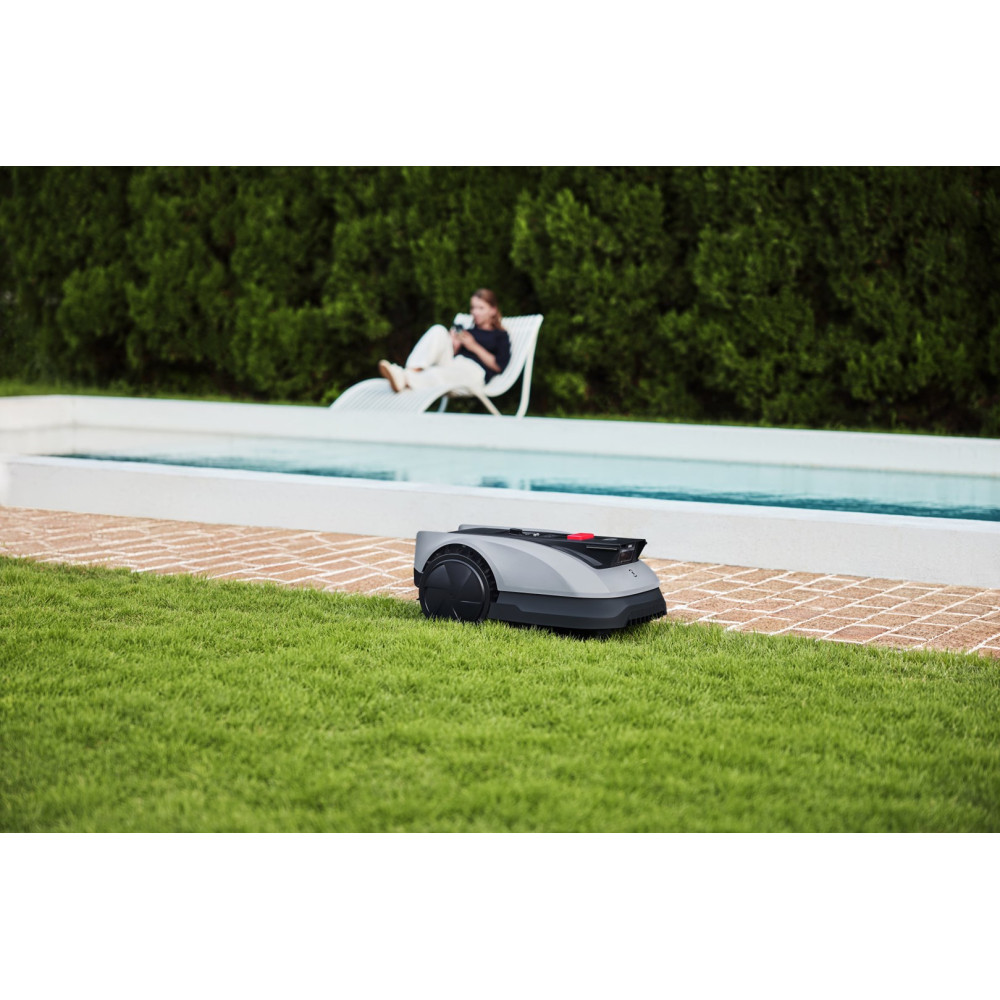 Robot koszący trawę na trawniku obok basenu, w tle osoba na leżaku.
