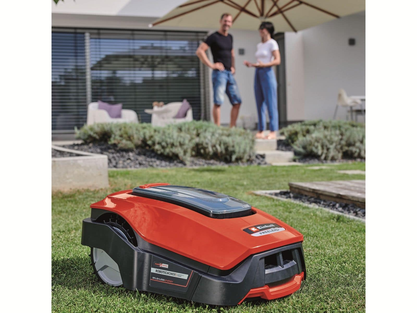 Czerwony robot koszący stoi na zielonym trawniku z parą w tle.