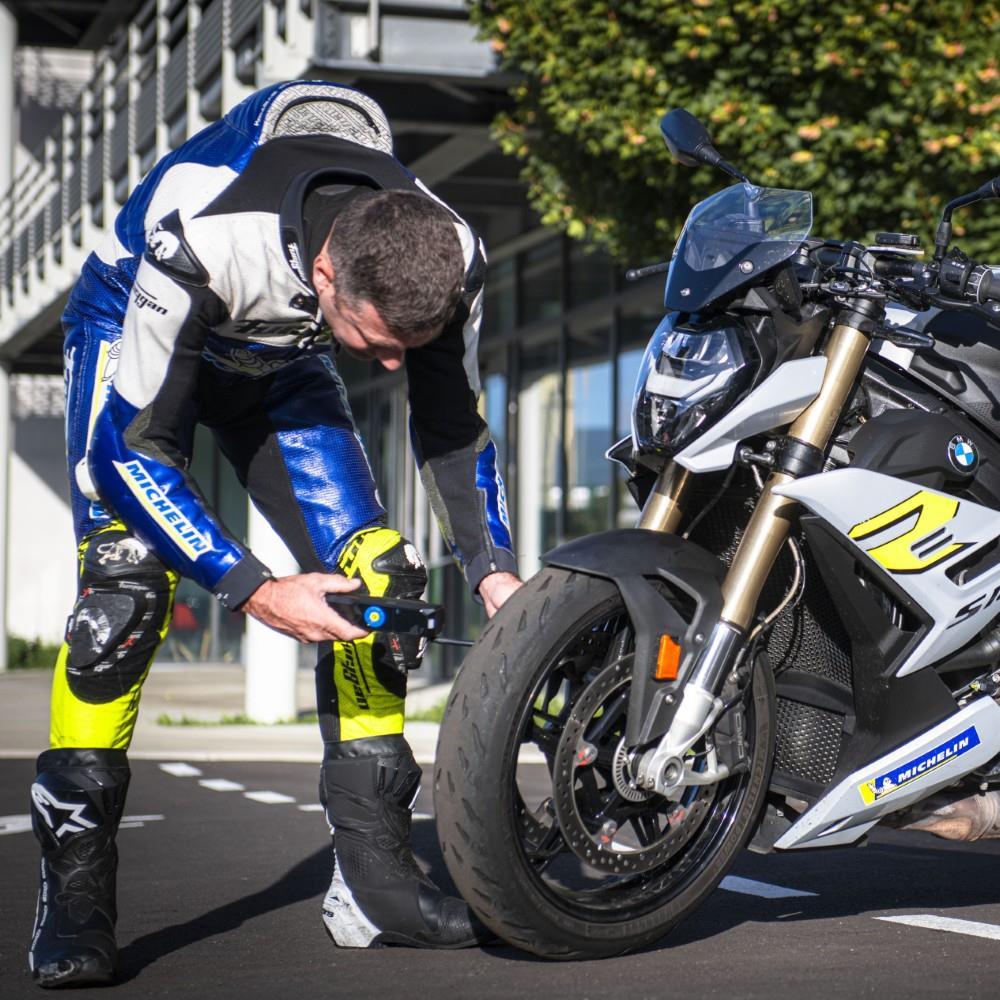 Mavi ve sarı motosiklet tulumu giymiş bir adam, beyaz bir BMW motosikletin lastiğini kontrol ediyor.