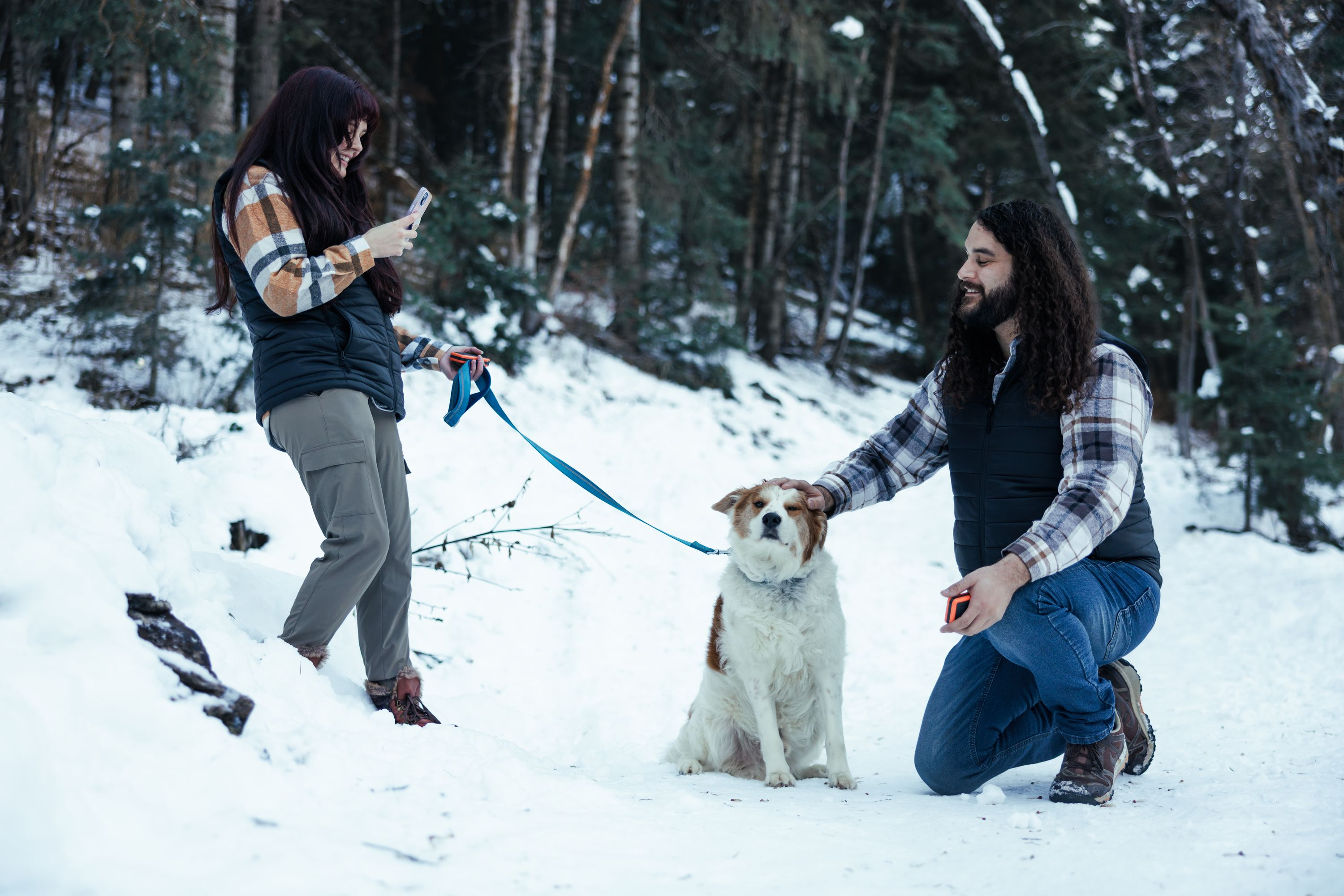 Ein Mann kniet bei einem Hund im Schnee, eine Frau steht daneben und schaut auf ihr Handy.