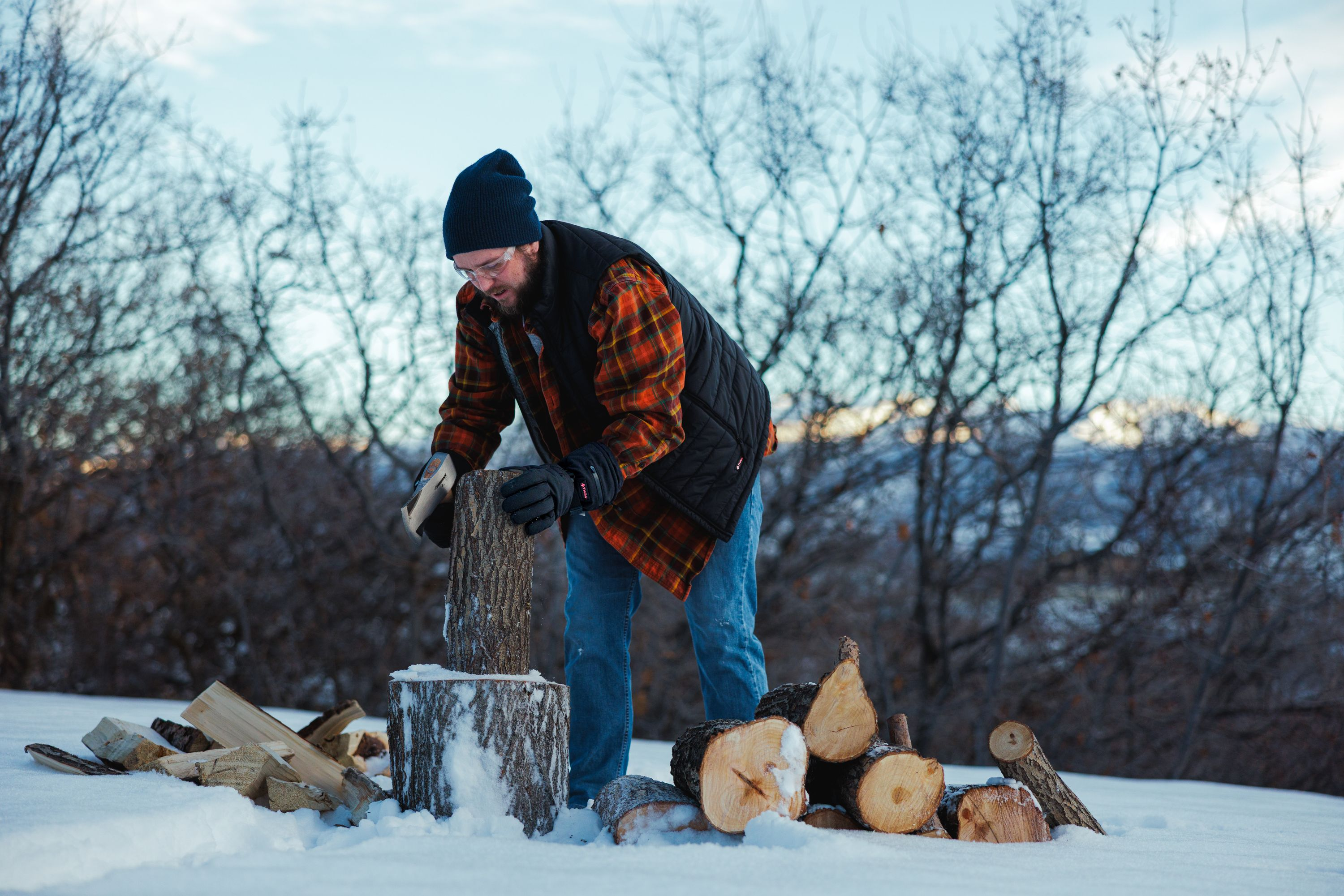 Mann spaltet Holz im Schnee mit Axt. Bäume im Hintergrund.