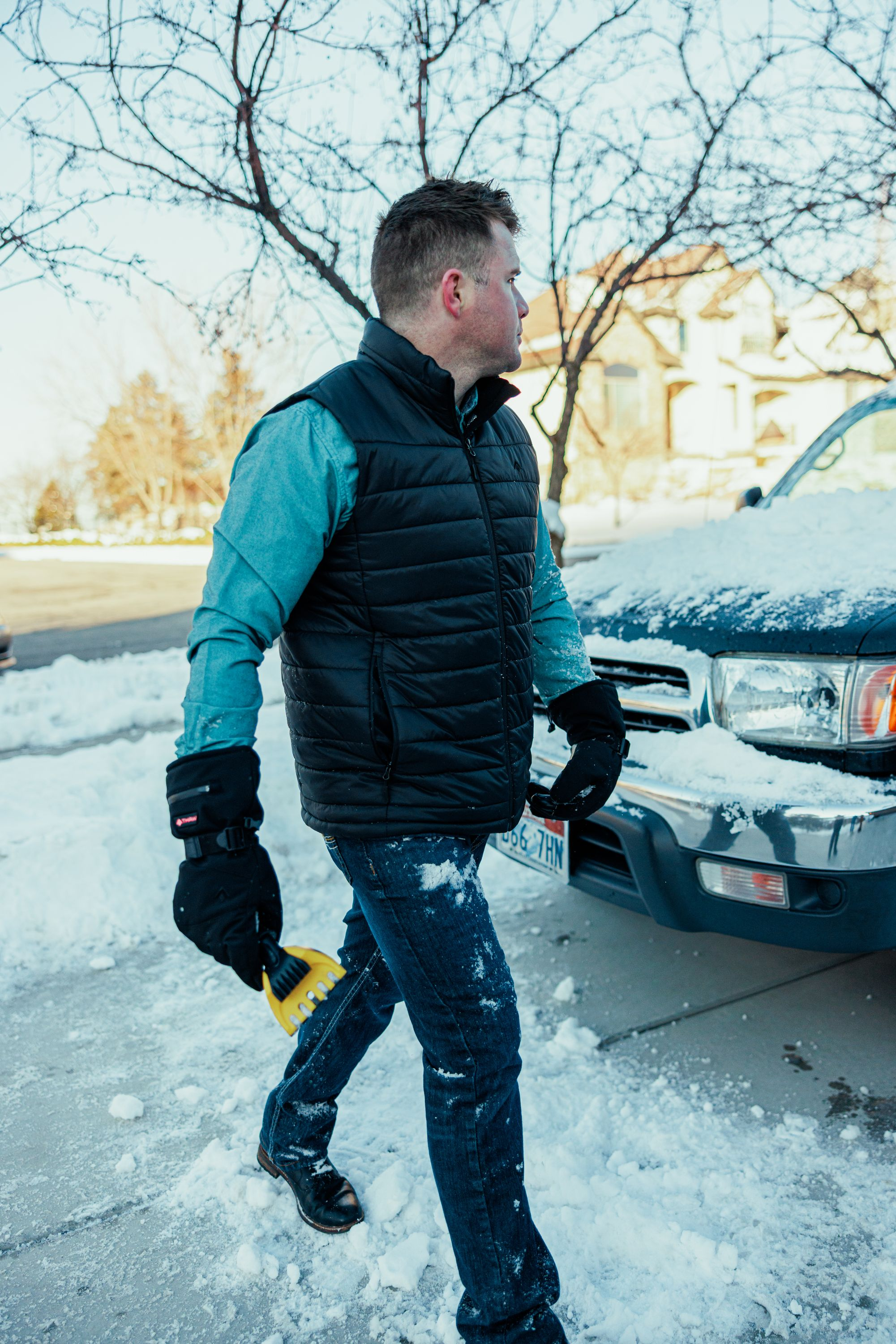 Ein Mann mit blauem Hemd und schwarzer Weste schabt Schnee vom Auto.