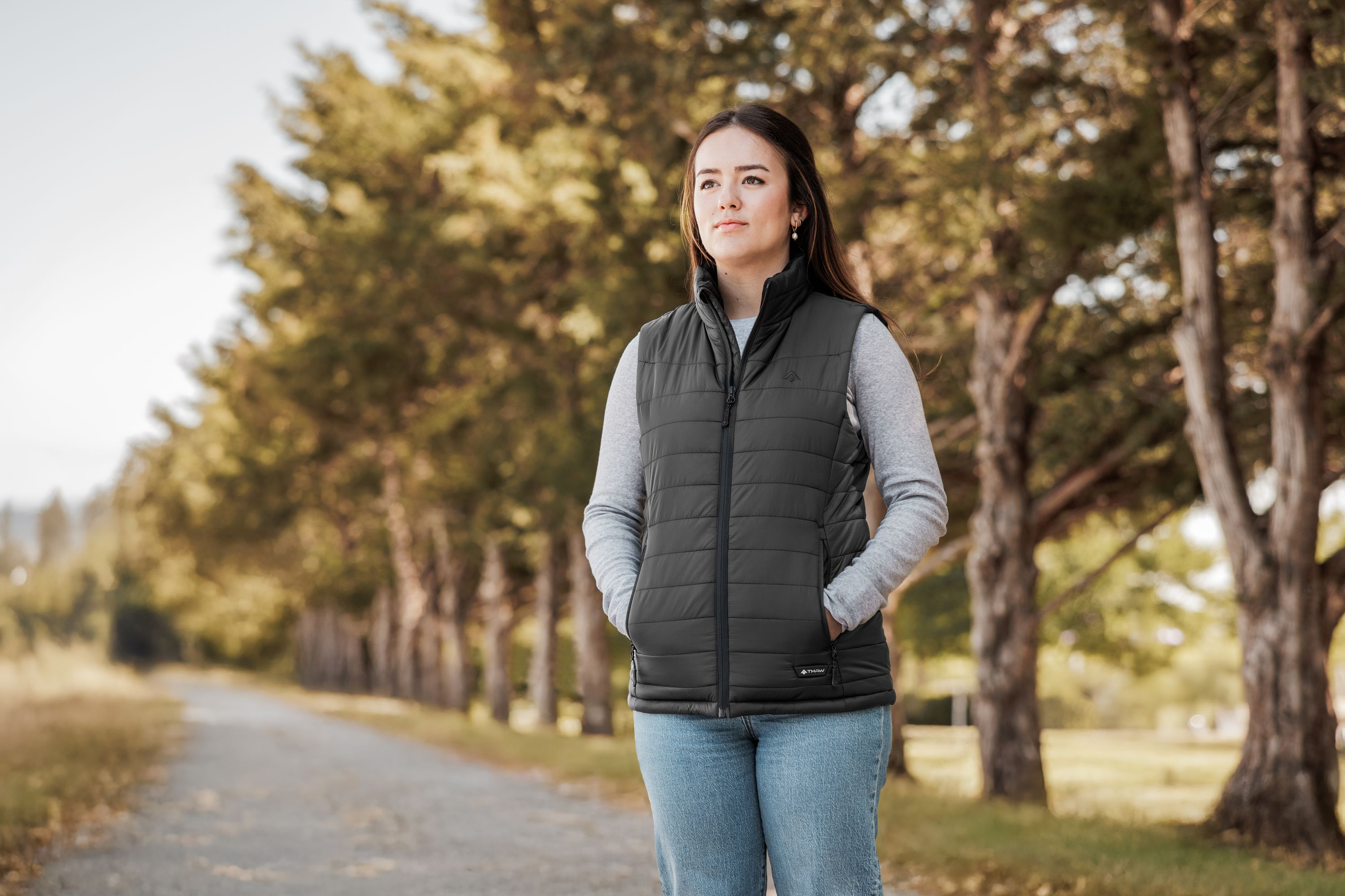 Eine Frau in einem grauen Pullover und schwarzer Weste steht auf einem von Bäumen gesäumten Weg.