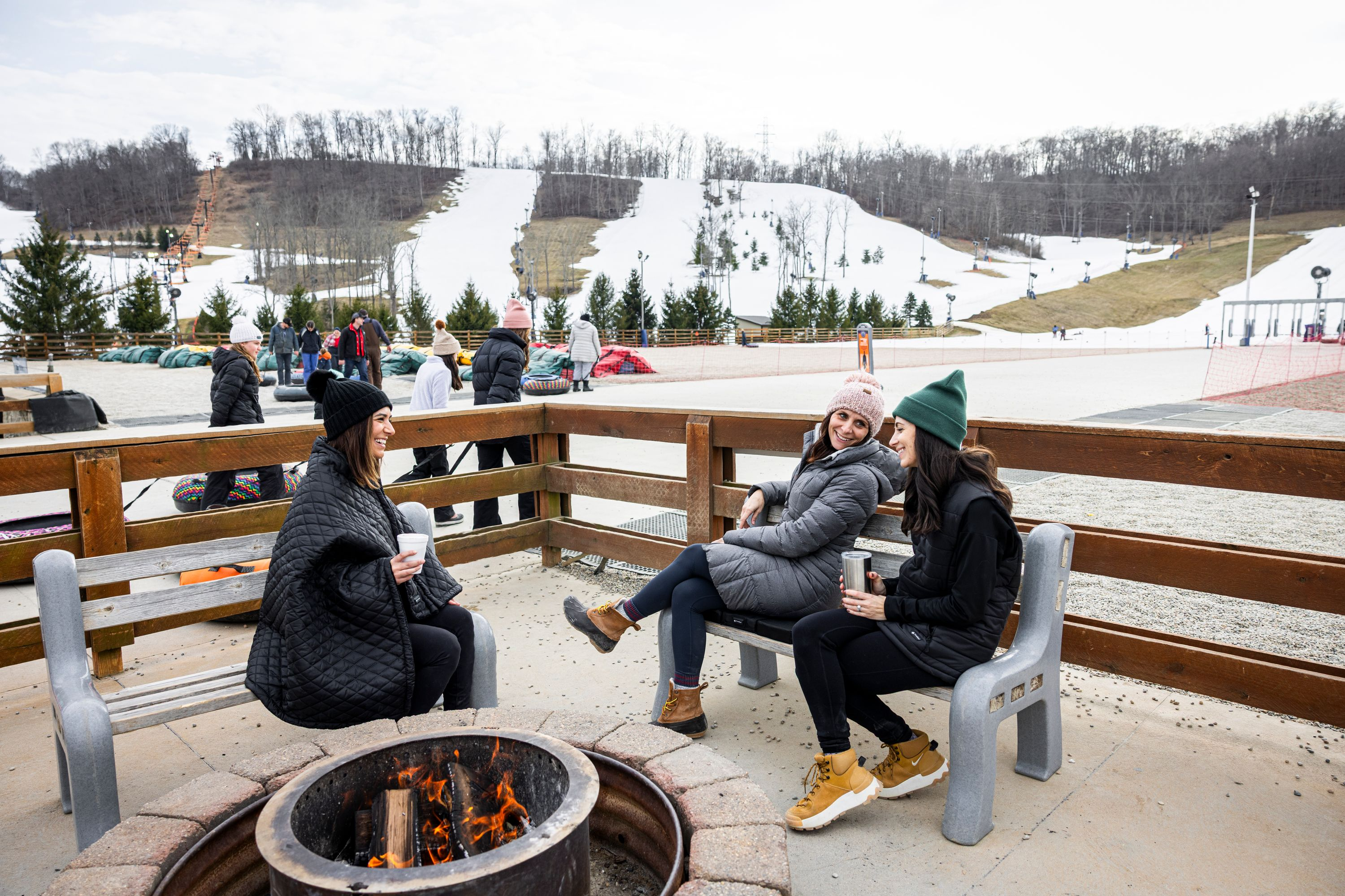 Leute sitzen an einem Feuerplatz mit einer Skipiste im Hintergrund.