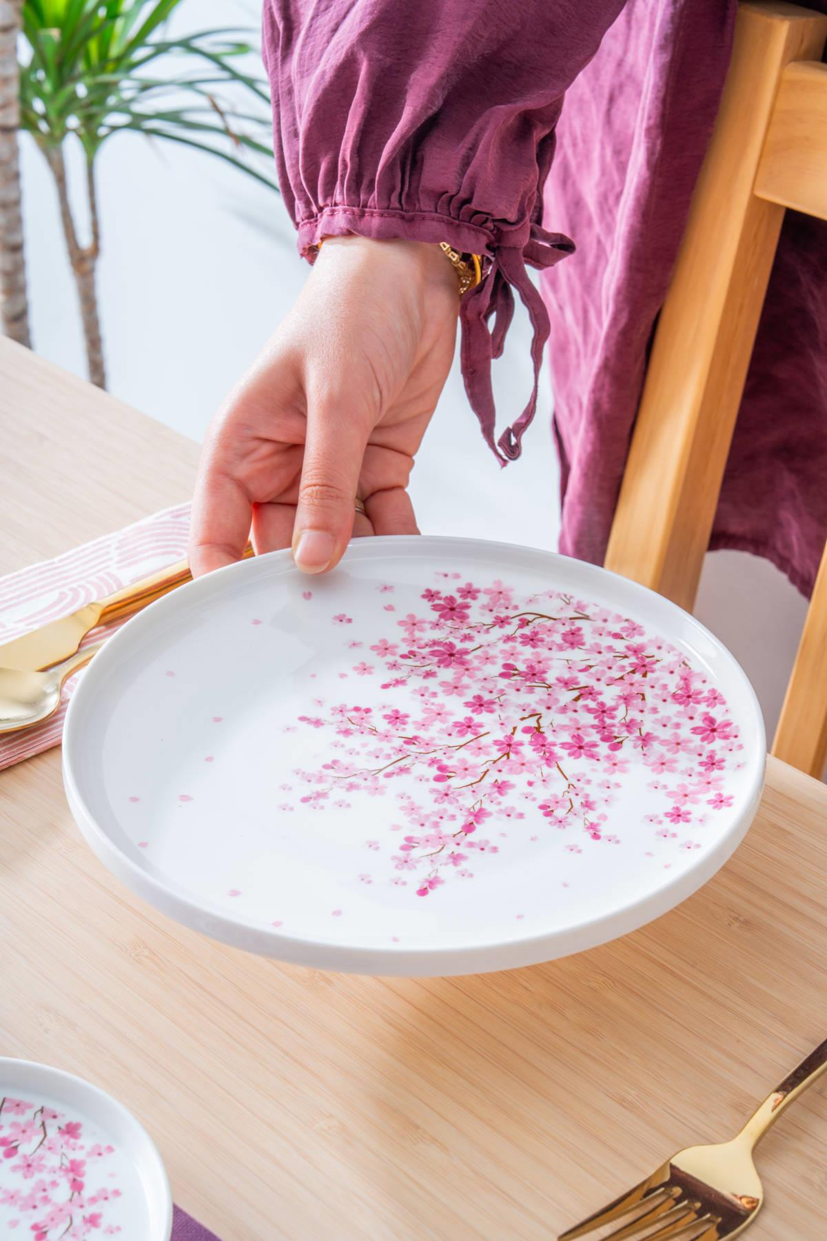 Bir el, ahşap masa üzerinde pembe kiraz çiçeği desenli beyaz bir tabak tutuyor.