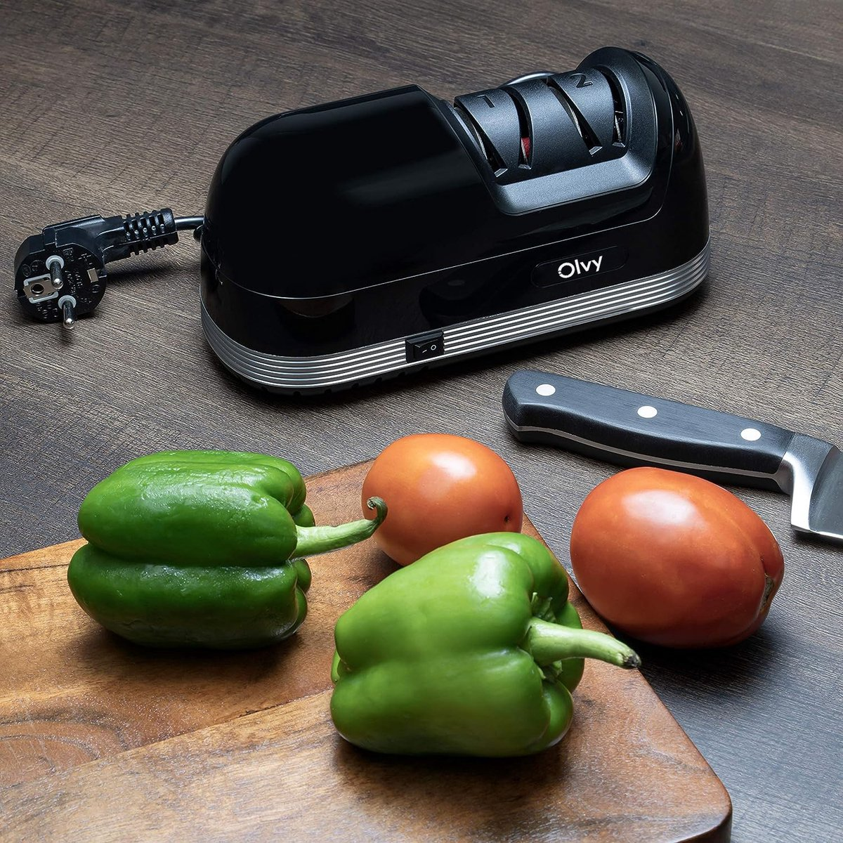 A black electric knife sharpener with cord, next to green peppers, tomatoes, and a knife.