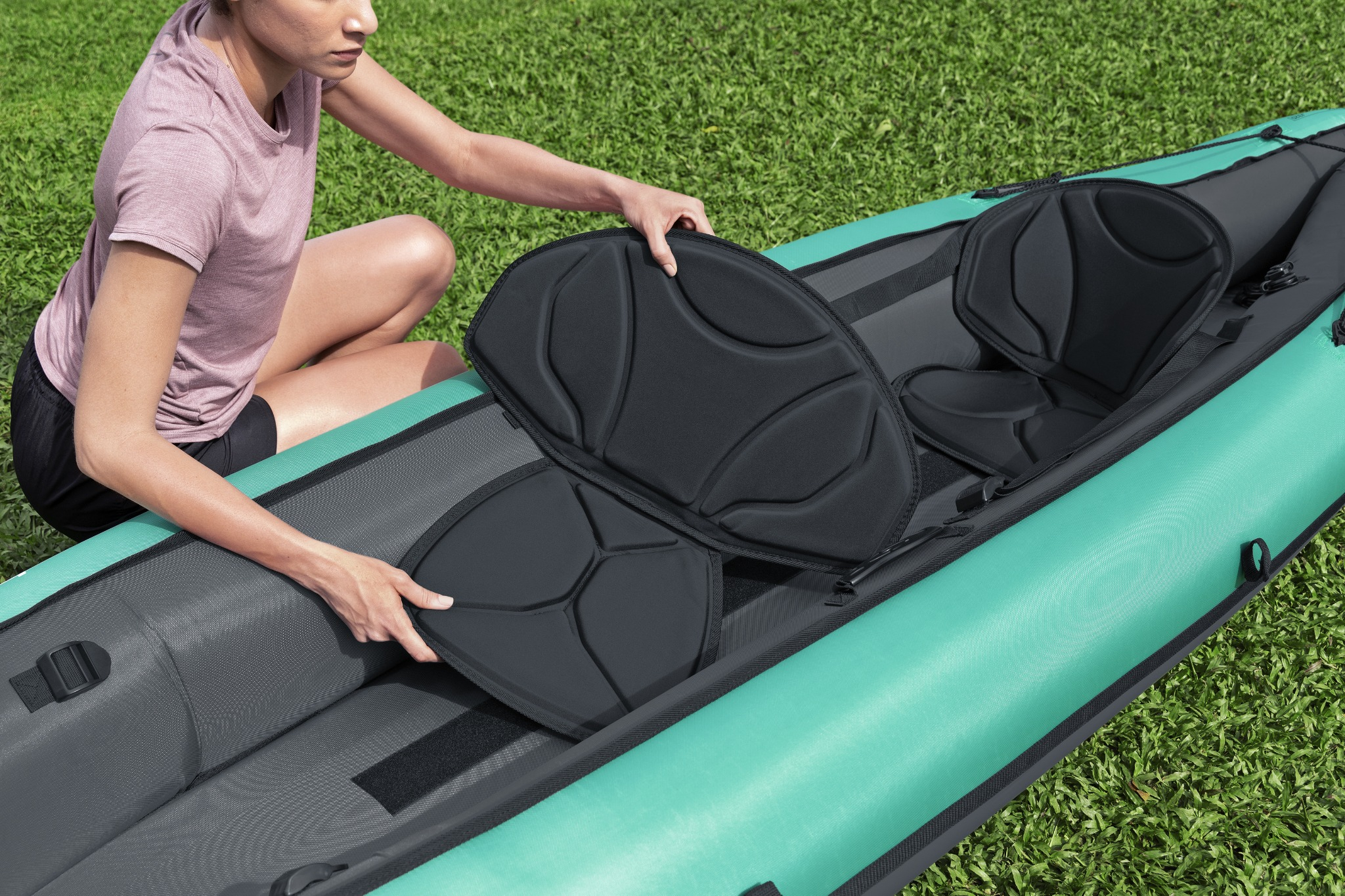 Mujer con camiseta rosa coloca asiento de kayak en kayak verde sobre césped.