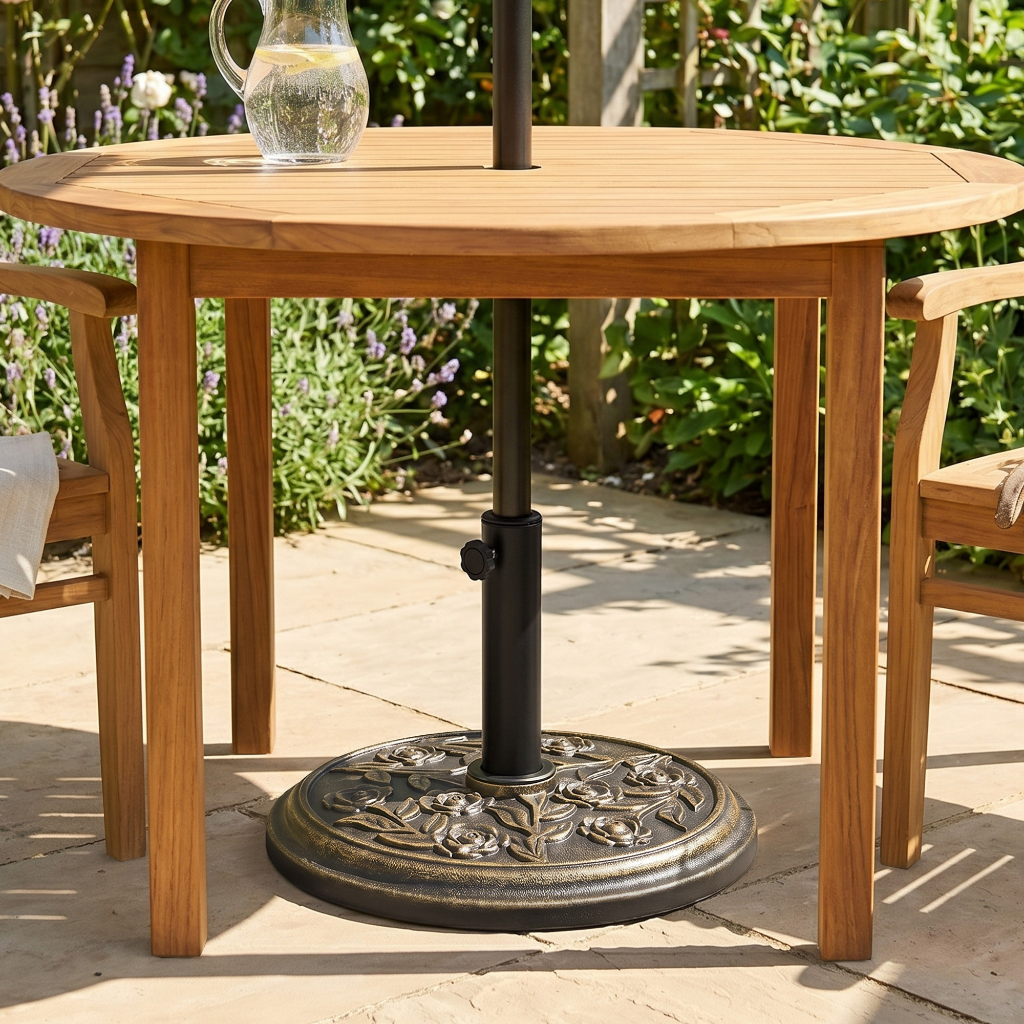 Una mesa redonda de madera con soporte para sombrilla y jarra de agua en un patio de piedra.