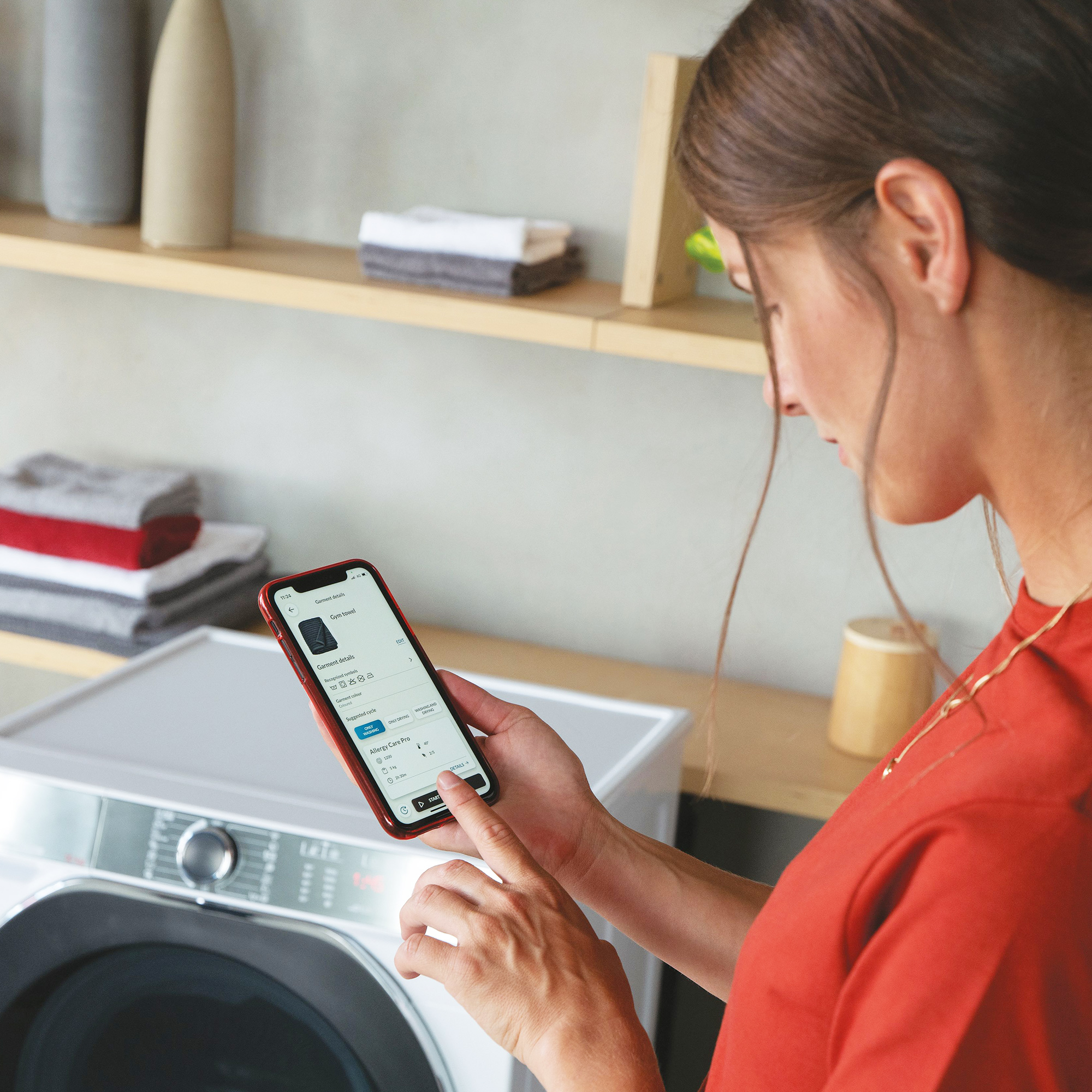 Eine Frau benutzt ihr Smartphone, um eine Waschmaschine zu steuern. Stapel von Handtüchern sind sichtbar.