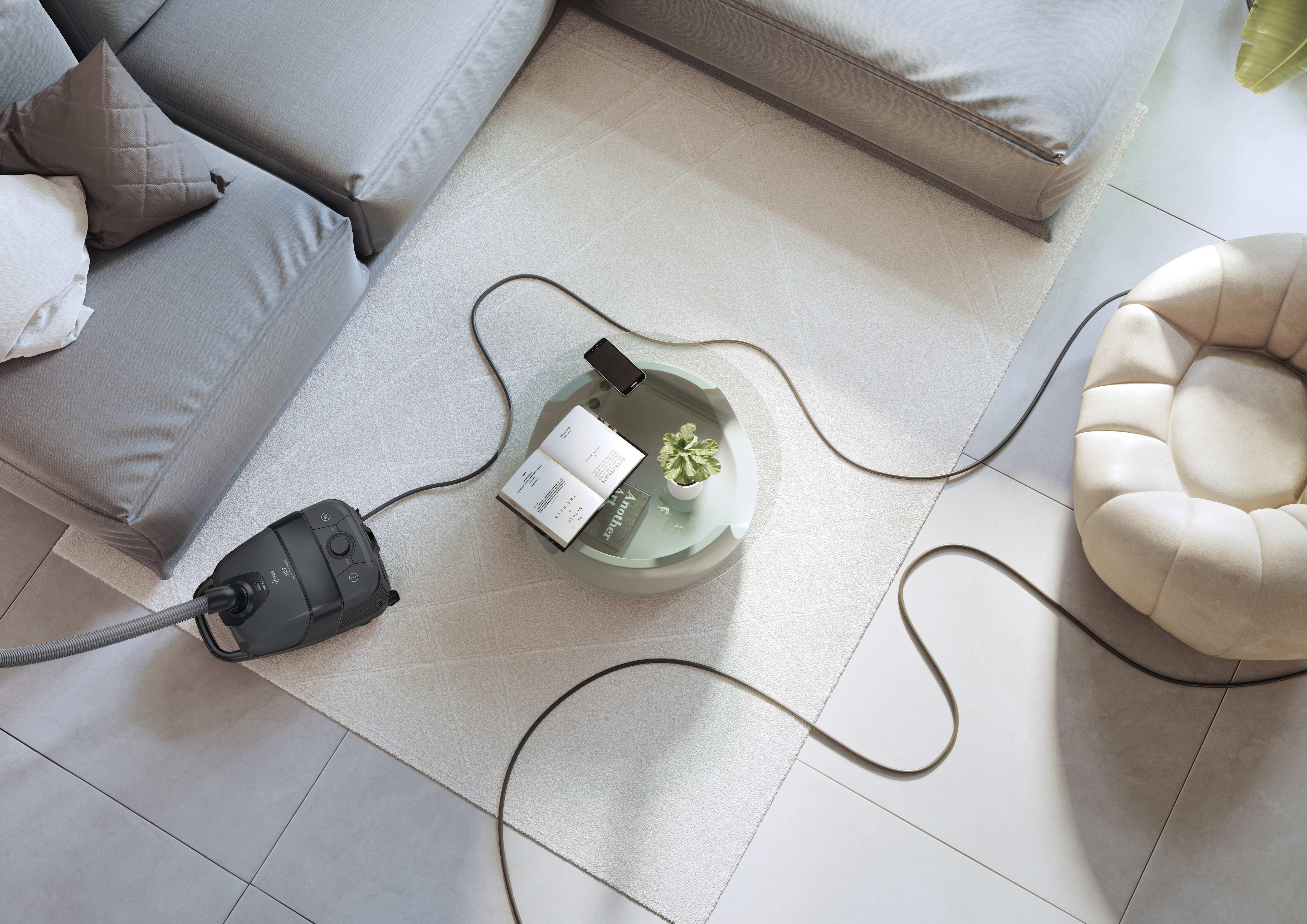 Ein grauer Staubsauger steht auf einem hellen Teppich neben einem Sofa.