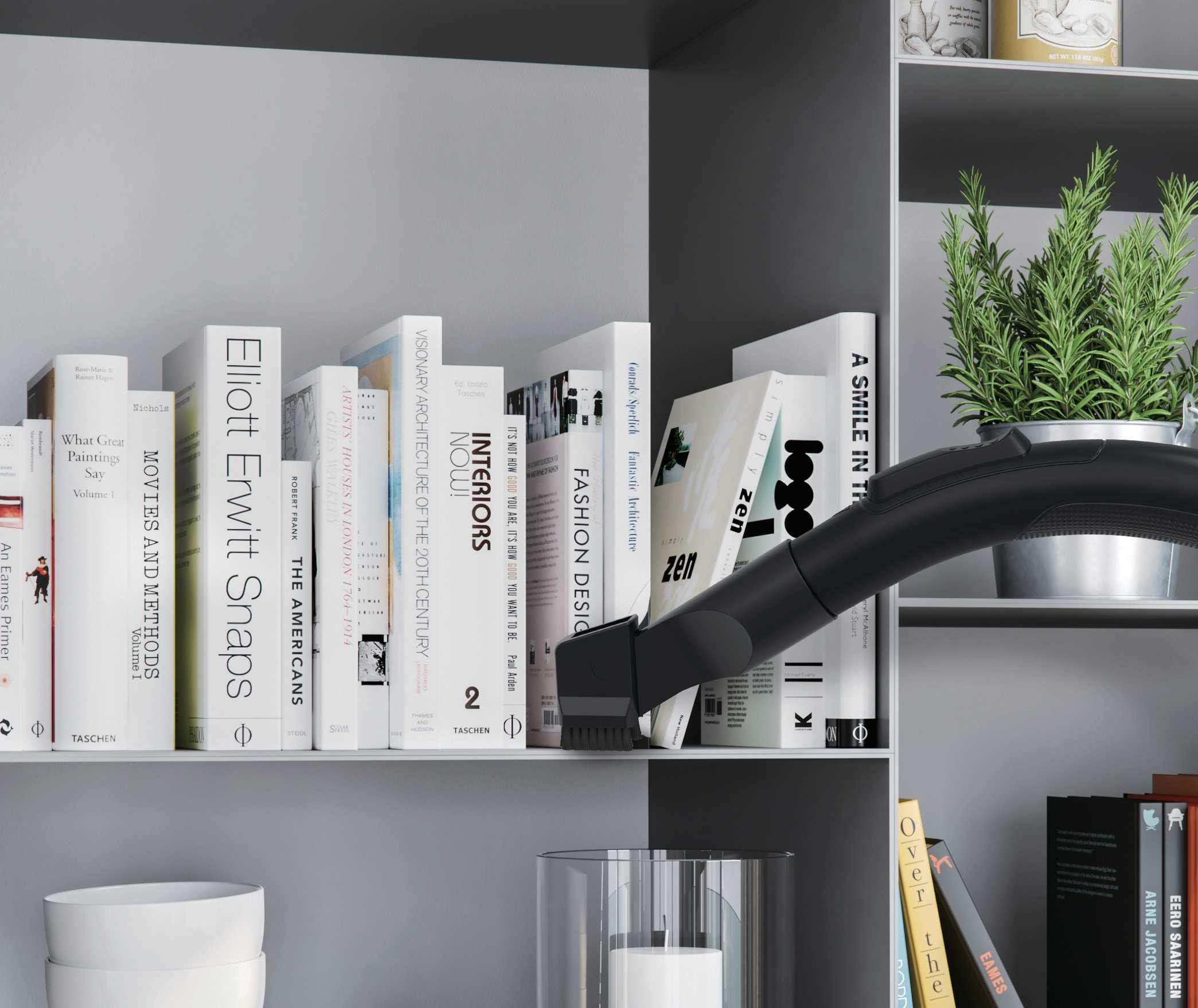 Ein Staubsaugeraufsatz reinigt ein Bücherregal mit Büchern und einer Topfpflanze.