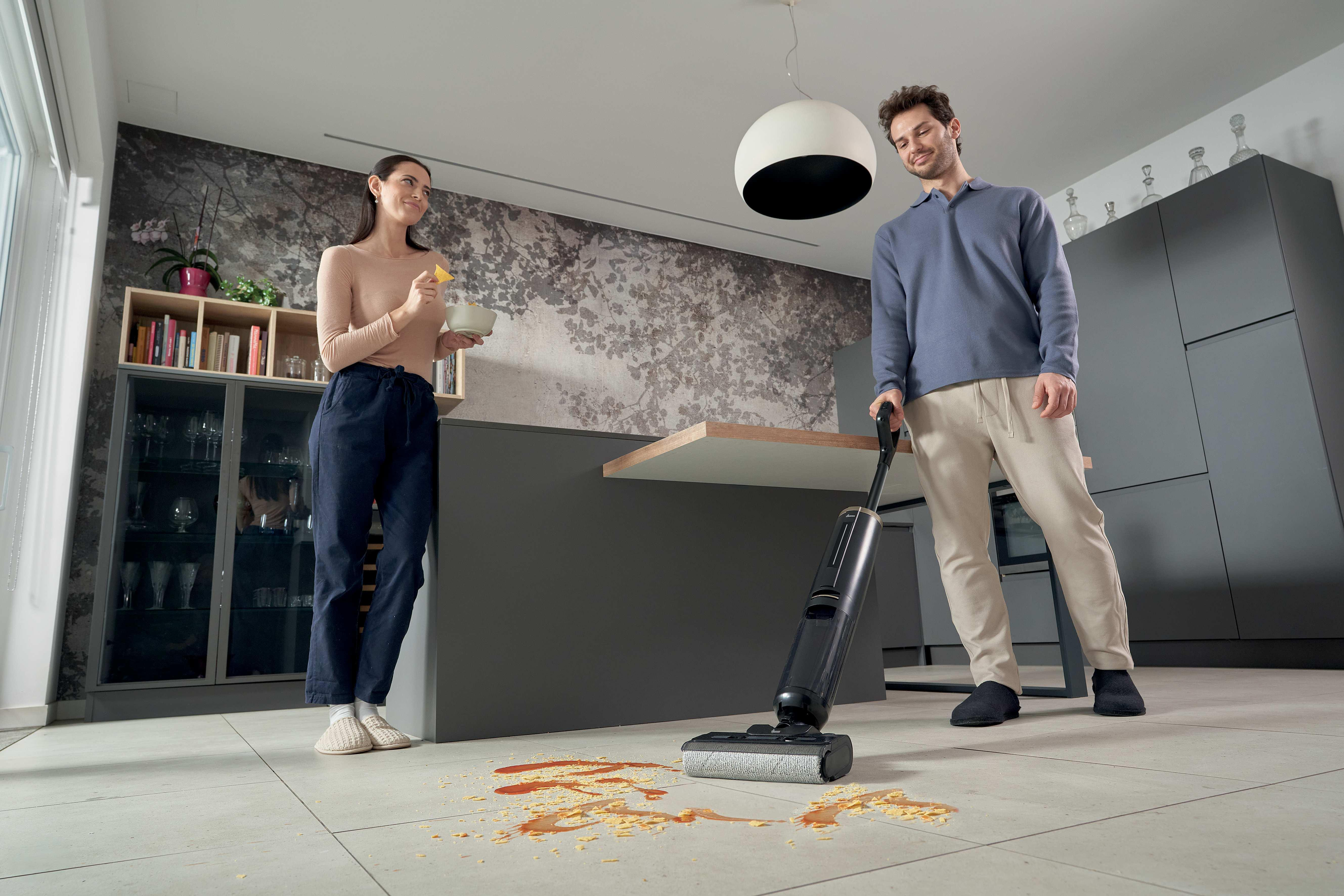 Homme passe l'aspirateur sur de la nourriture renversée, femme mange des chips.