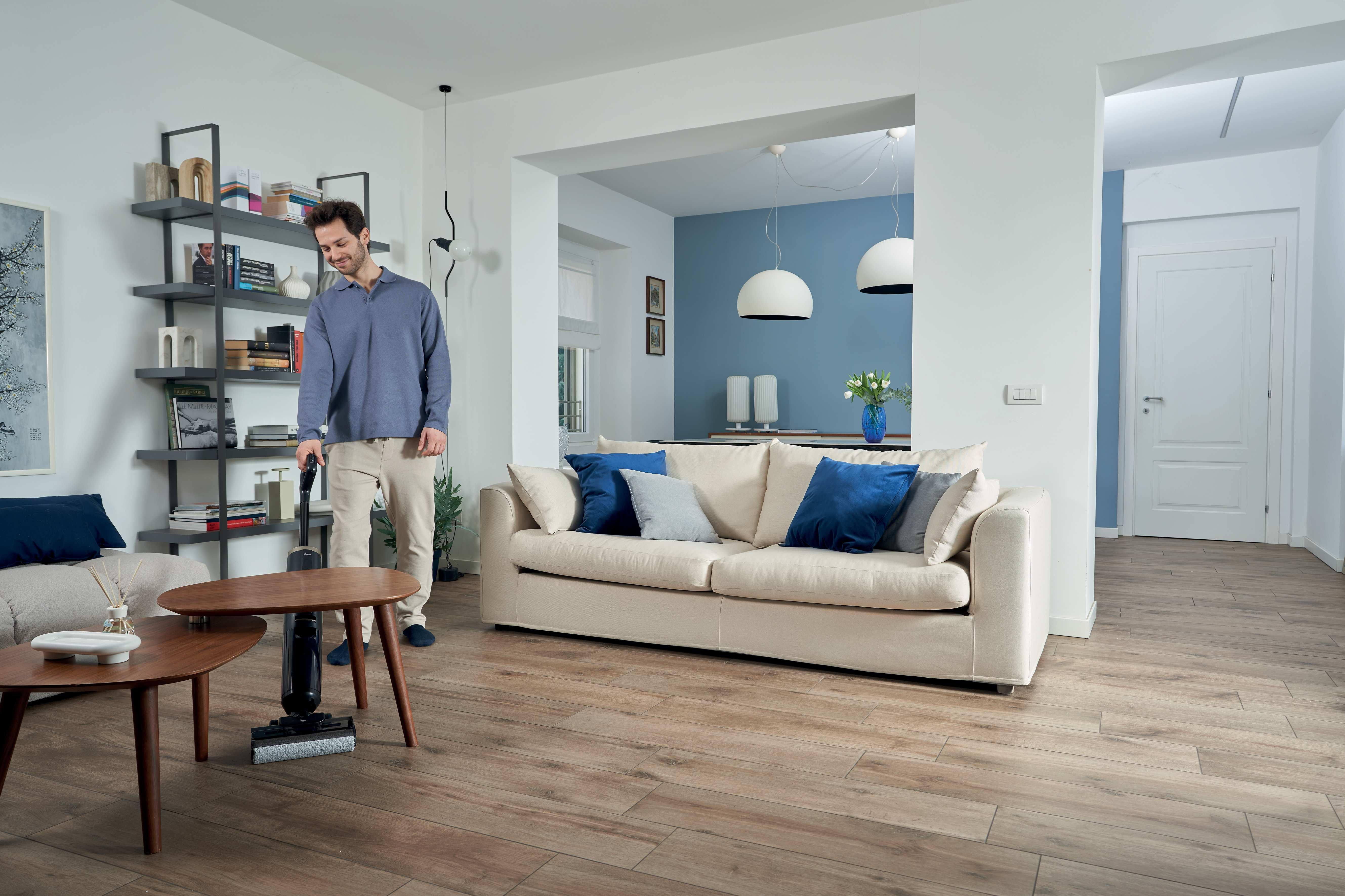 Un homme passe l'aspirateur sur un parquet dans un salon moderne avec canapé et étagères.