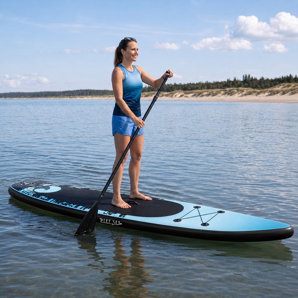 Frau paddelt auf einem blau-schwarzen SUP-Board auf ruhigem Wasser unter blauem Himmel.
